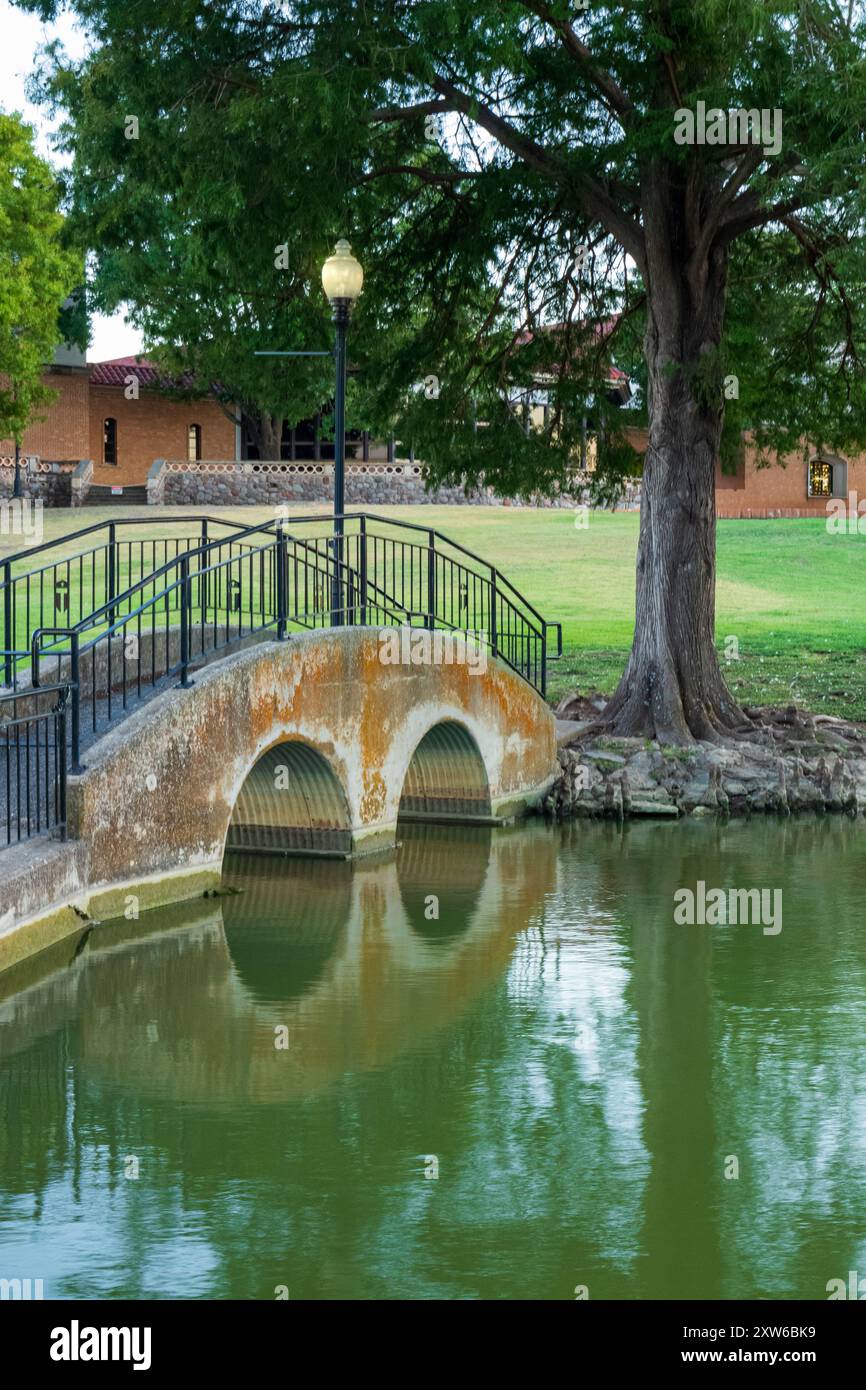 Ruhige Fußgängerbrücke über einen grünen Teich mit Bäumen, Eisengeländern und einem Steingebäude im Hintergrund, das in einem ruhigen Park liegt. Stockfoto