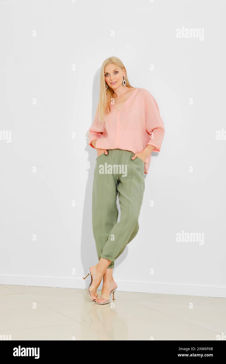 Die blonde Frau in Bluse und Hose mit V-Ausschnitt hält die Hände in den Taschen und lehnt sich an die weiße Wand Stockfoto