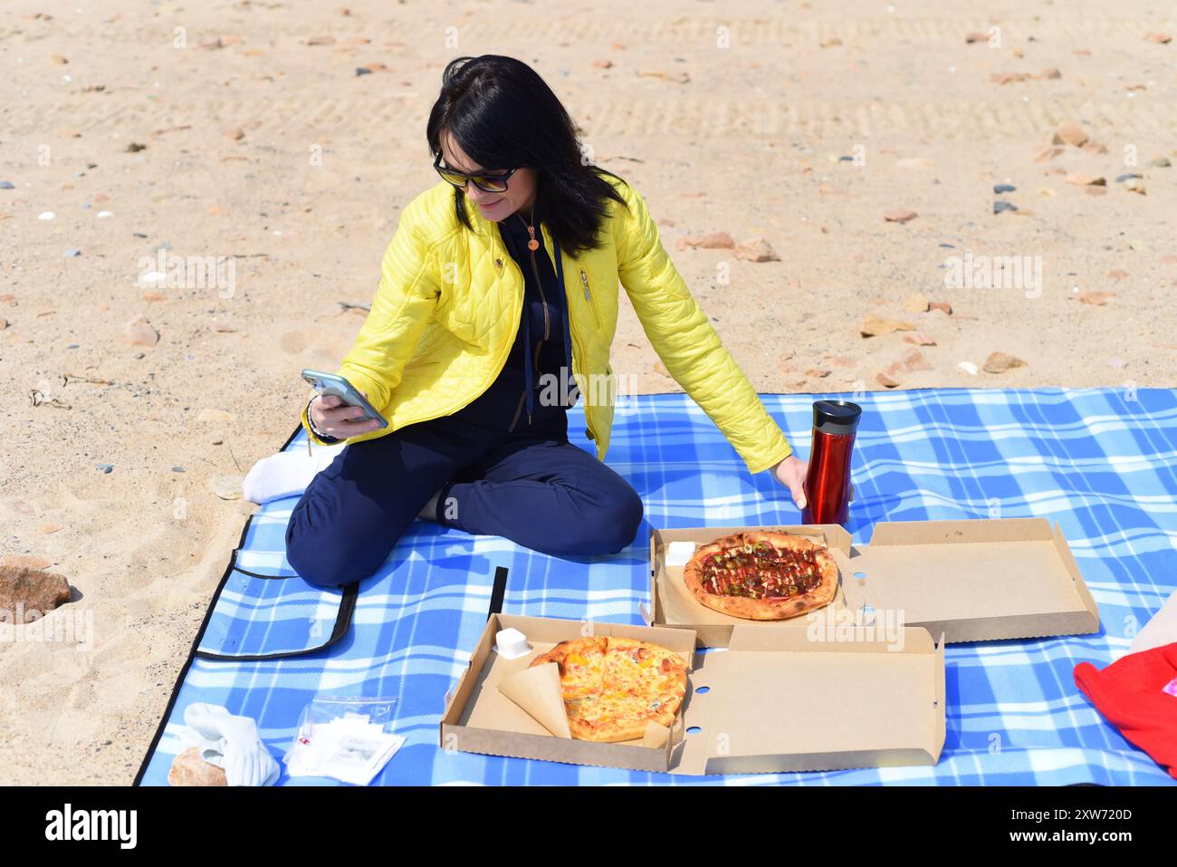 Russische 45-jährige Frau, die Pizza essen will, während sie am Telefon scrollt und am Strand sitzt Stockfoto