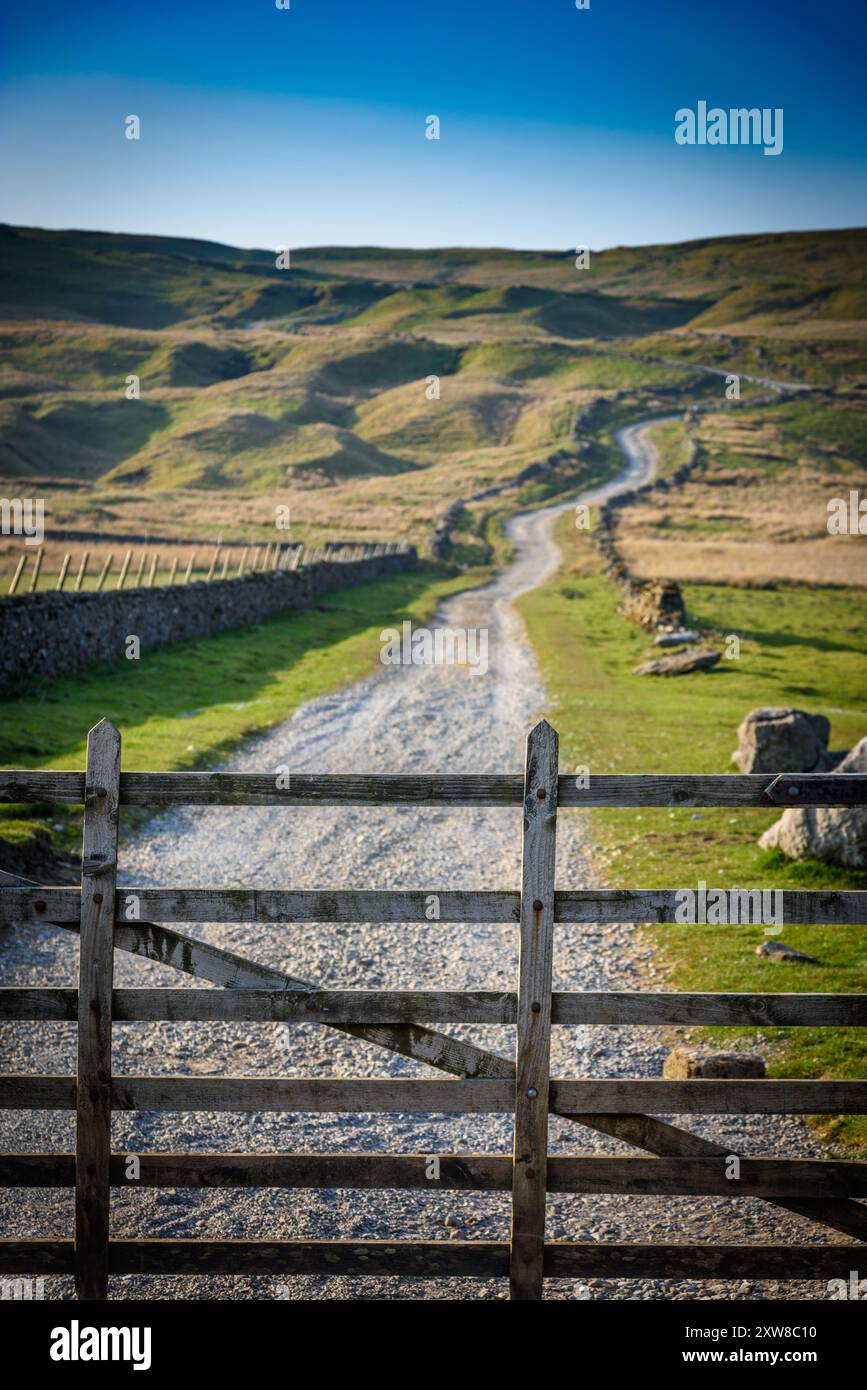 Ein hölzernes Tor steht offen und offenbart einen Schotterweg, der durch sanfte grüne und braune Hügel unter einem klaren blauen Himmel hinaufsteigt. Üppiges Gras grenzt an die p Stockfoto