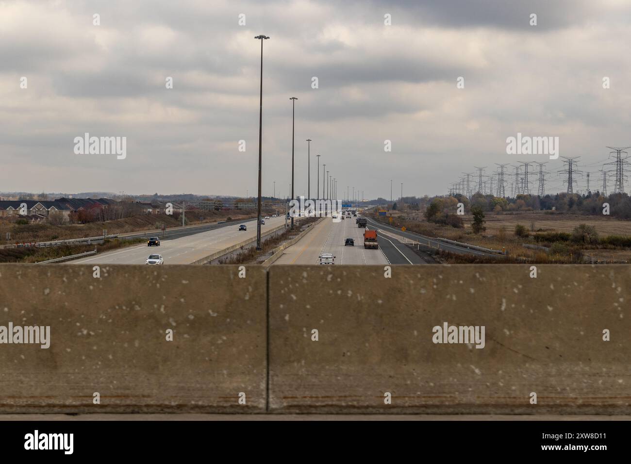 Über einer belebten Autobahn, die von Stromleitungen und entfernten Gebäuden flankiert wird, erhebt sich ein bedeckter Himmel, der über einer Betonbarriere betrachtet wird. Aufgenommen in Toronto, Kanada. Stockfoto