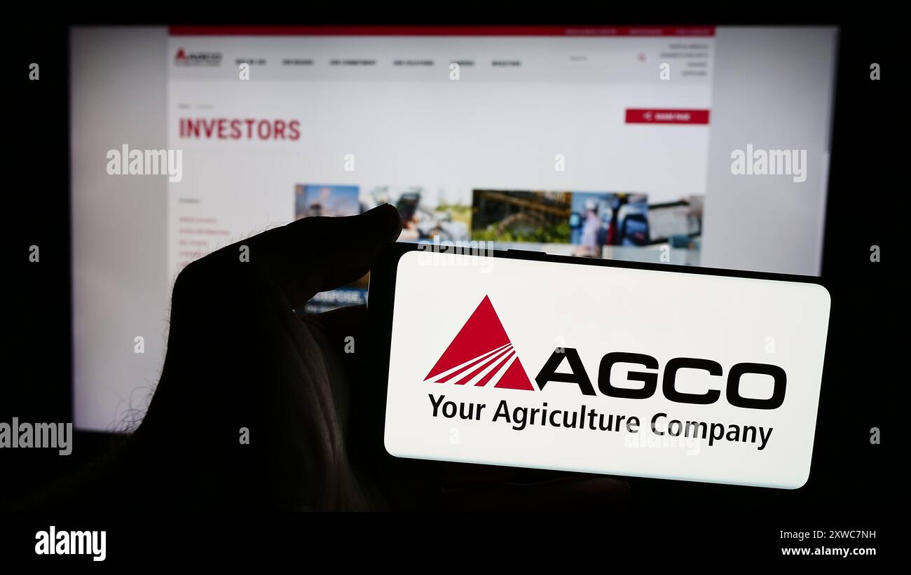 Person mit Smartphone und Logo des US-amerikanischen Landmaschinenunternehmens AGCO Corporation vor der Website. Schwerpunkt auf dem Display des Telefons. Stockfoto