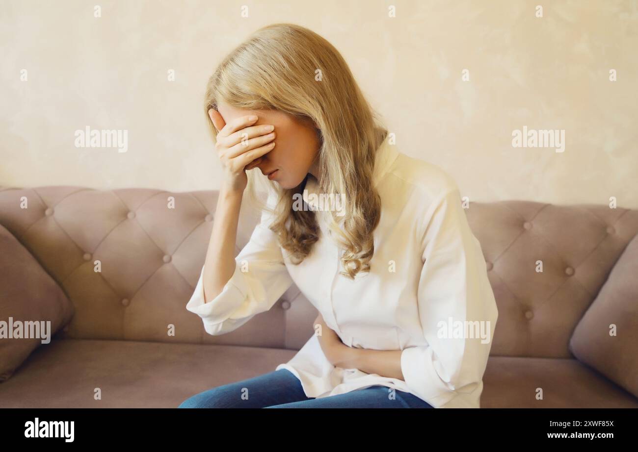 Junge Frau hat Hände auf den Bauch und leidet unter Bauchschmerzen, Bauchschmerzen, fühlen sich schlecht mit Schmerzen, die auf einer Couch zu Hause sitzen Stockfoto