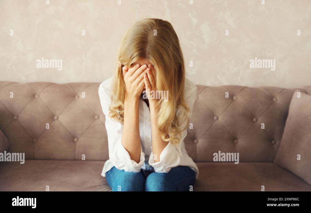 Verärgerte traurige, weinende Frau, die ihre Augen und ihr Gesicht mit den Händen bedeckt, während sie geistiges oder körperliches Leid erlebt, Ermüdung auf der Couch im Zimmer sitzt Stockfoto