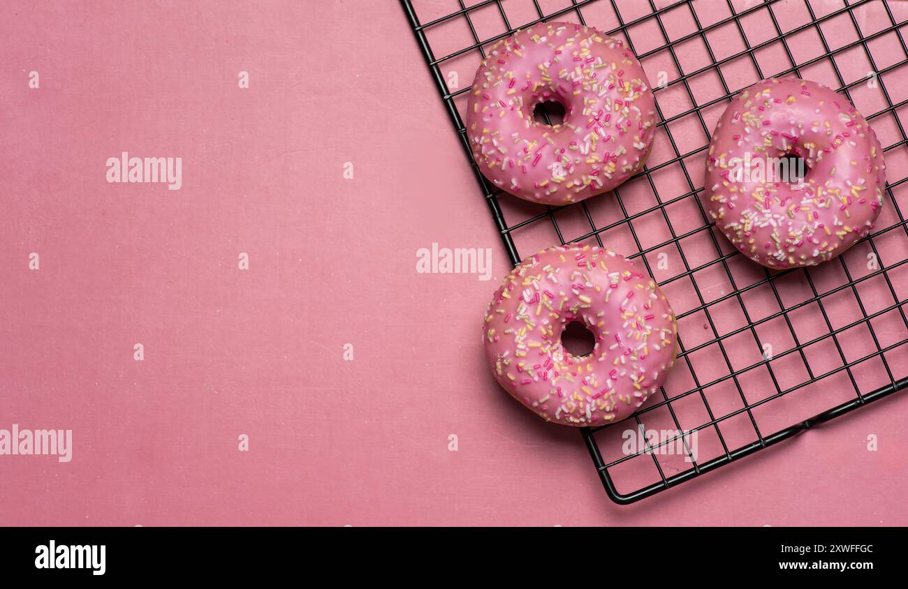 Drei rosafarbene Donuts mit bunten Streuseln ruhen teilweise auf einem Kühlregal auf einem rosafarbenen Hintergrund und strahlen ein einladendes und köstliches aus Stockfoto