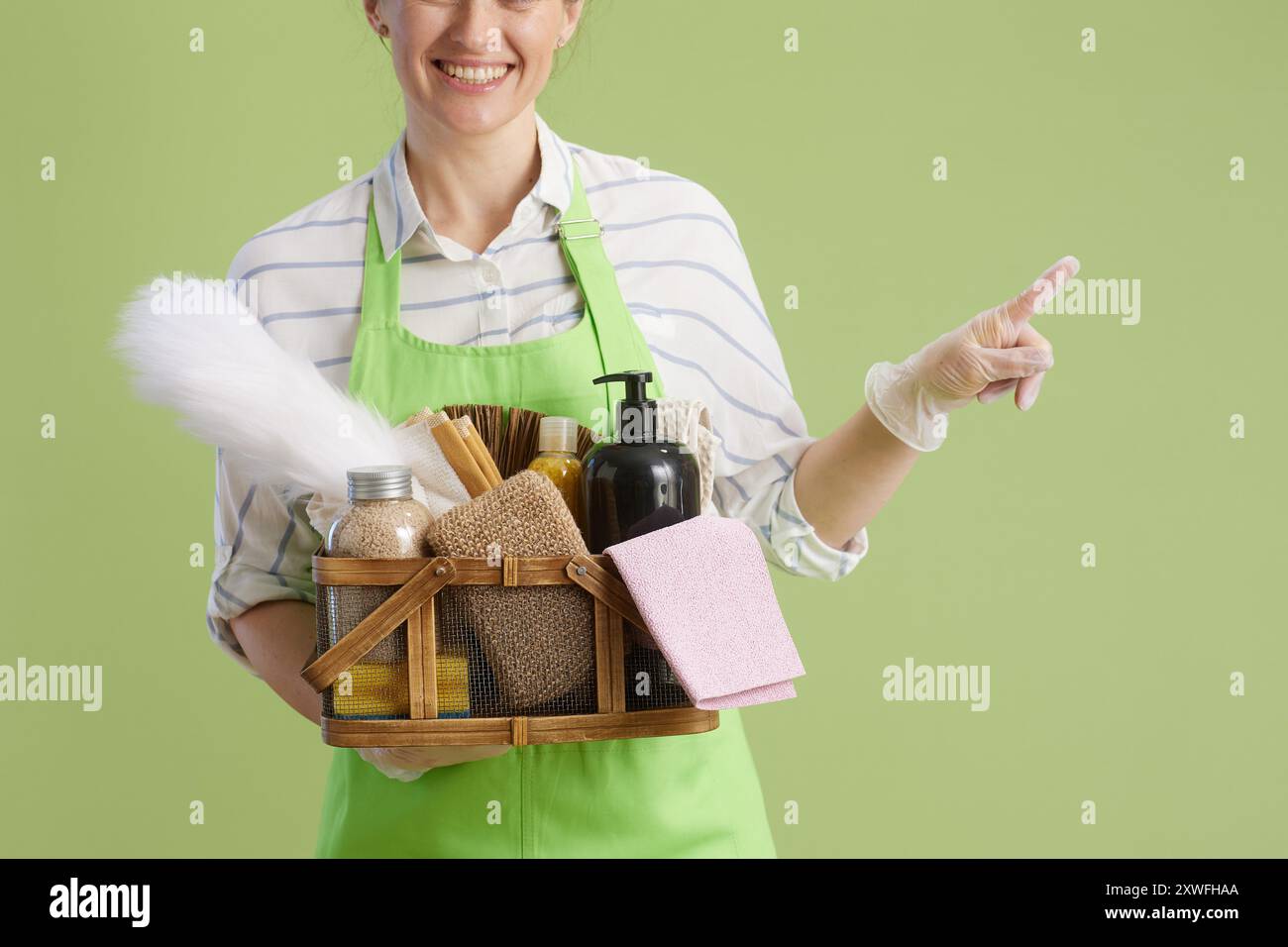 Frühjahrsputz. Nahaufnahme über lächelnde moderne Hausfrau mittleren Alters in grüner Schürze und Gummihandschuhe isoliert auf grünem Hintergrund mit Korb von Öko Clea Stockfoto