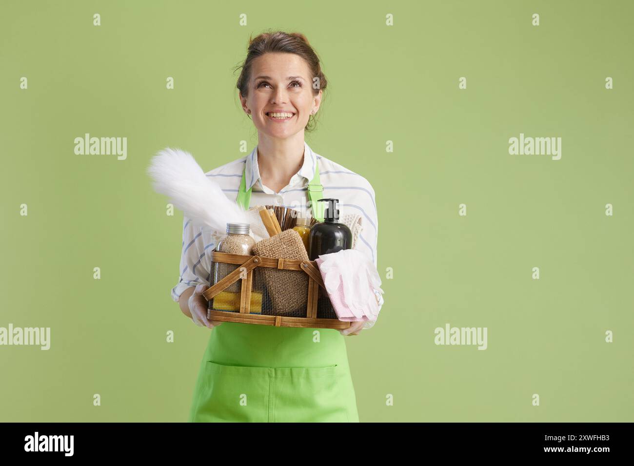 Frühjahrsputz. Glückliche moderne 40 Jahre alte Hausfrau in grüner Schürze und Gummihandschuhen auf grünem Hintergrund mit Korb mit Öko-Reinigungsprodukten schauen Stockfoto