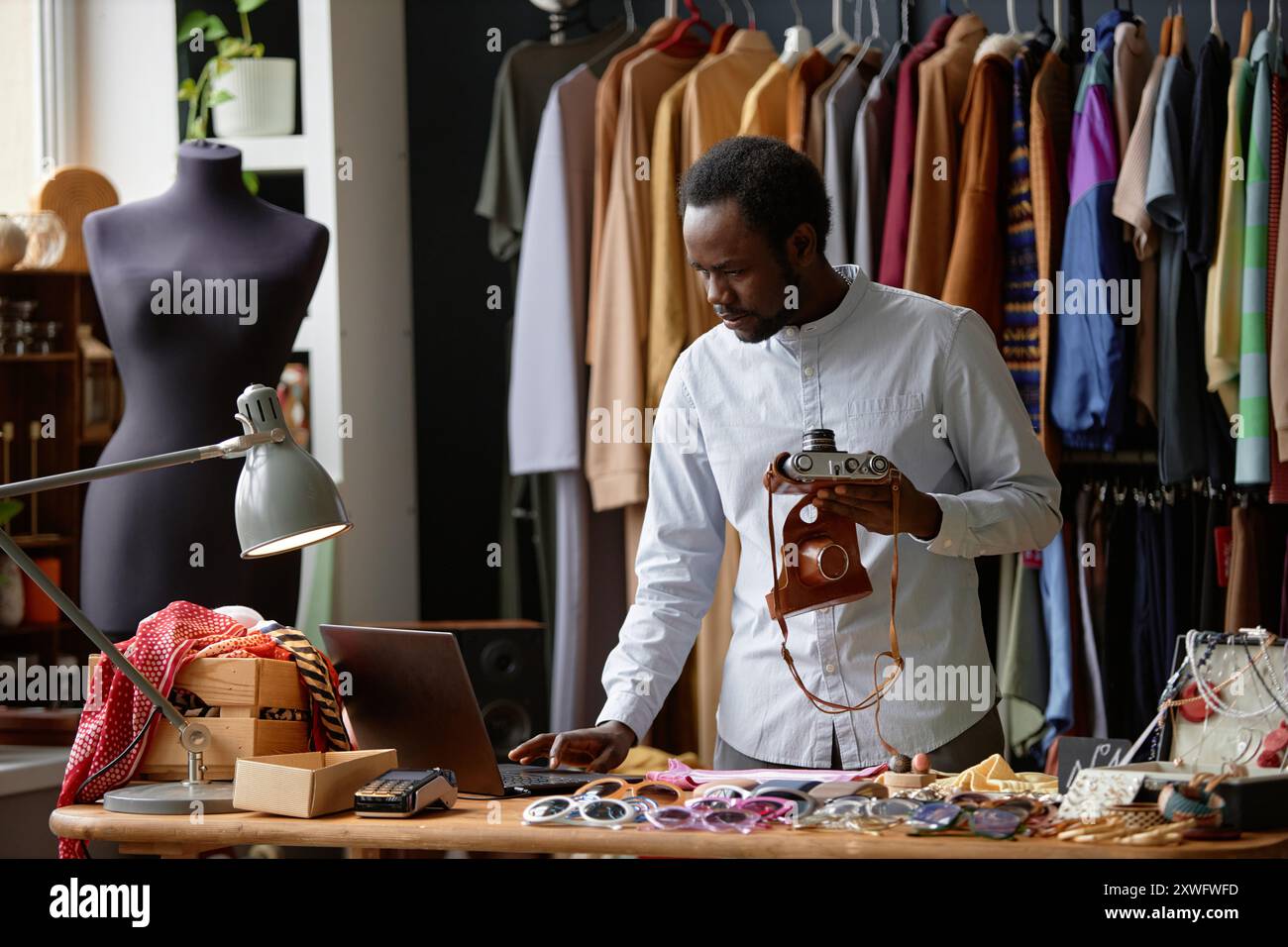 Männlicher afroamerikanischer Second-Hand-Ladenassistent in elegantem Hemd mit Laptop, der eine alte Fotokamera zum Verkauf bringt, während er in sozialen Netzwerken im Second-Hand-Shop, Kopierraum, aufnimmt Stockfoto