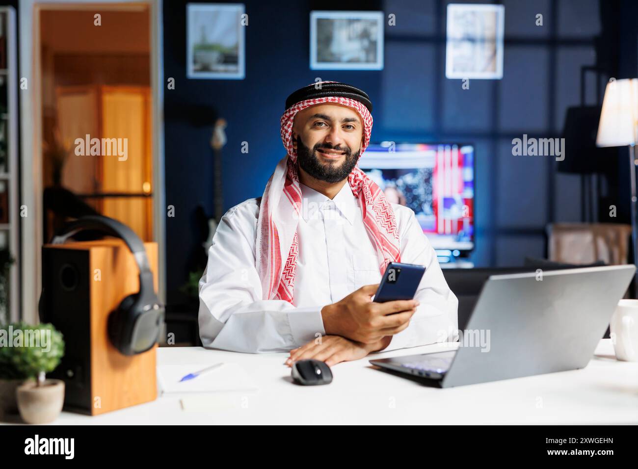 Ein lächelnder Geschäftsmann aus dem Nahen Osten, traditionell gekleidet, sitzt und hält sein Smartphone an einem Schreibtisch in seinem Büro. Ein arabischer Kerl greift ein Mobilgerät, sieht in die Kamera, mit einem Laptop in der Nähe. Stockfoto