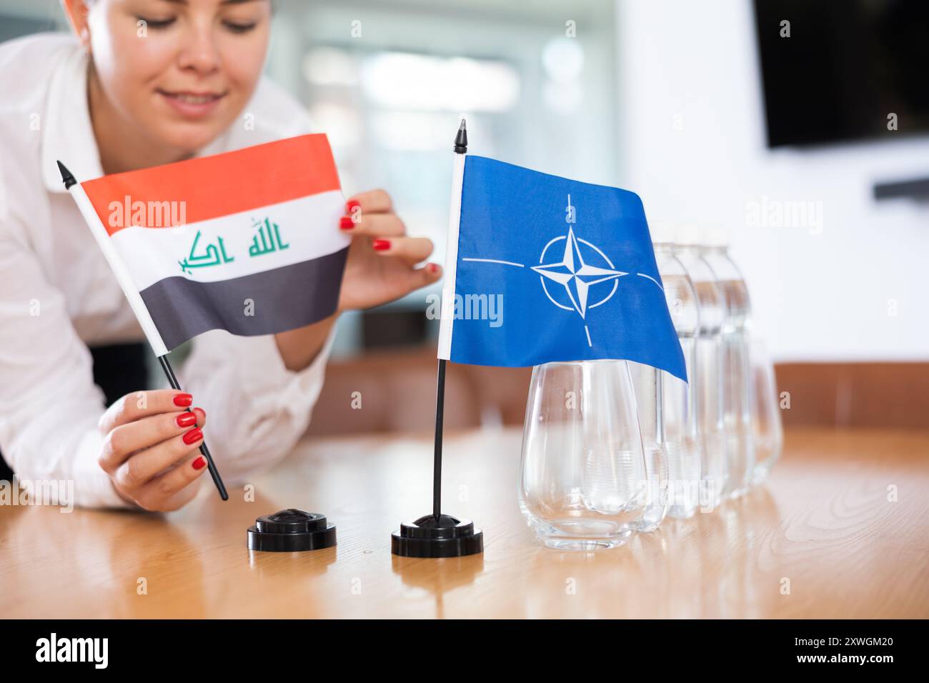 Frau Sekretärin bereitet ein Büro für Verhandlungen vor - sie legt Flaggen der NATO und des Irak Tisch Stockfoto