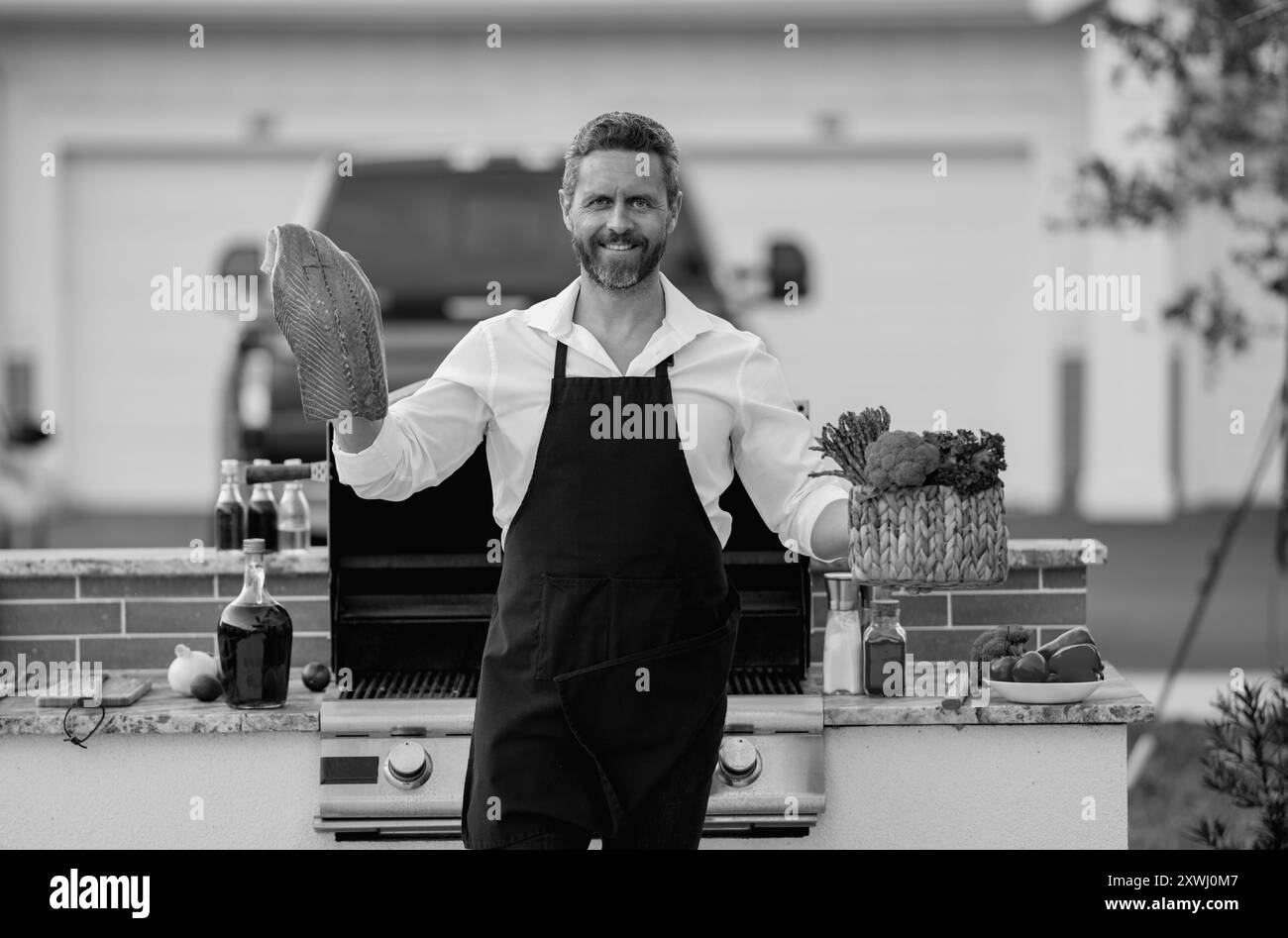 Mann im Koch Schürze kocht Lachsfisch auf Barbecue im Hinterhof des Hauses. Ein gutaussehender Mann, der Lachs-Barbecue zubereitet. Barbecue-Chef-Meister. Grill Stockfoto