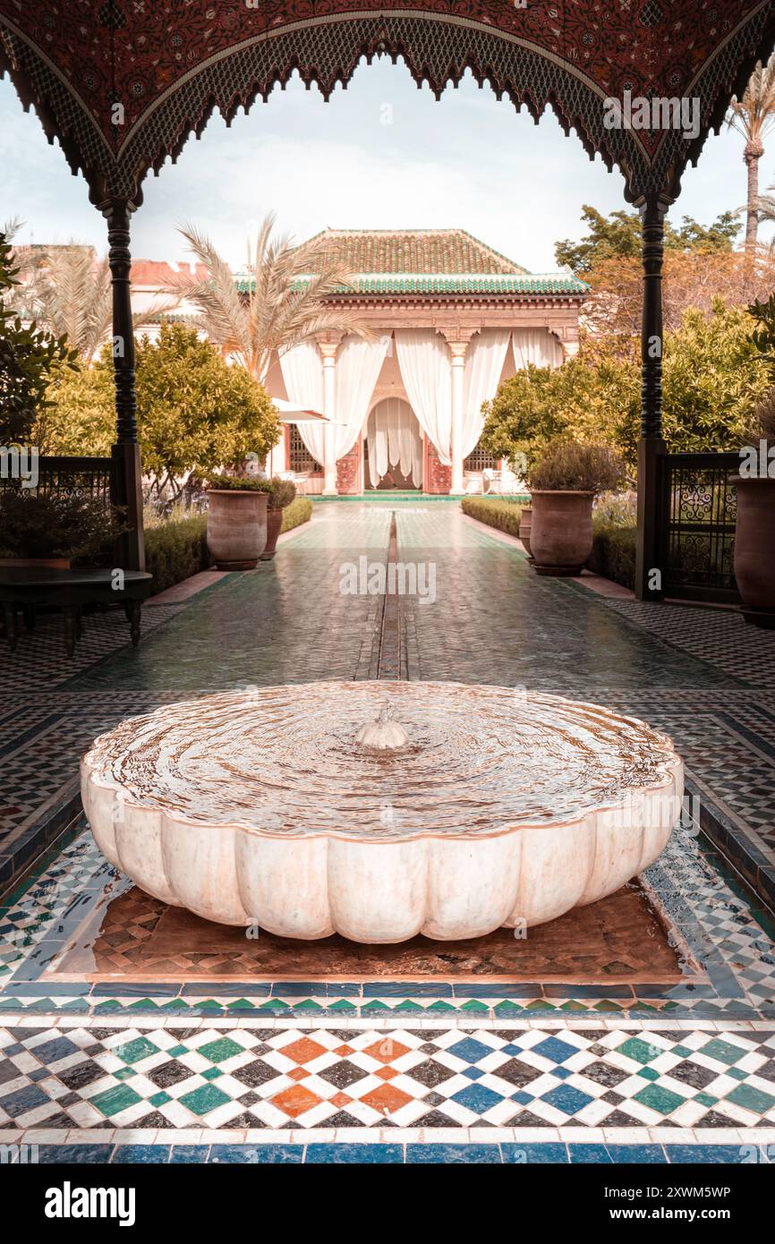 Ein ruhiger Innenhof mit einem großen, runden Brunnen, umgeben von komplizierten Fliesen und üppigem Grün. Der Hintergrund zeigt elegante drapierte Vorhänge Stockfoto