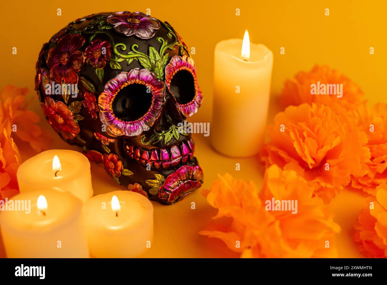 Ein schwarzer Schädel mit Blumendekorationen in Metallic-Optik, umgeben von Ringelblumen und Kerzen vor orangefarbenem Hintergrund. Eine lebhafte Hommage an Mexikos Día de los Muertos Stockfoto