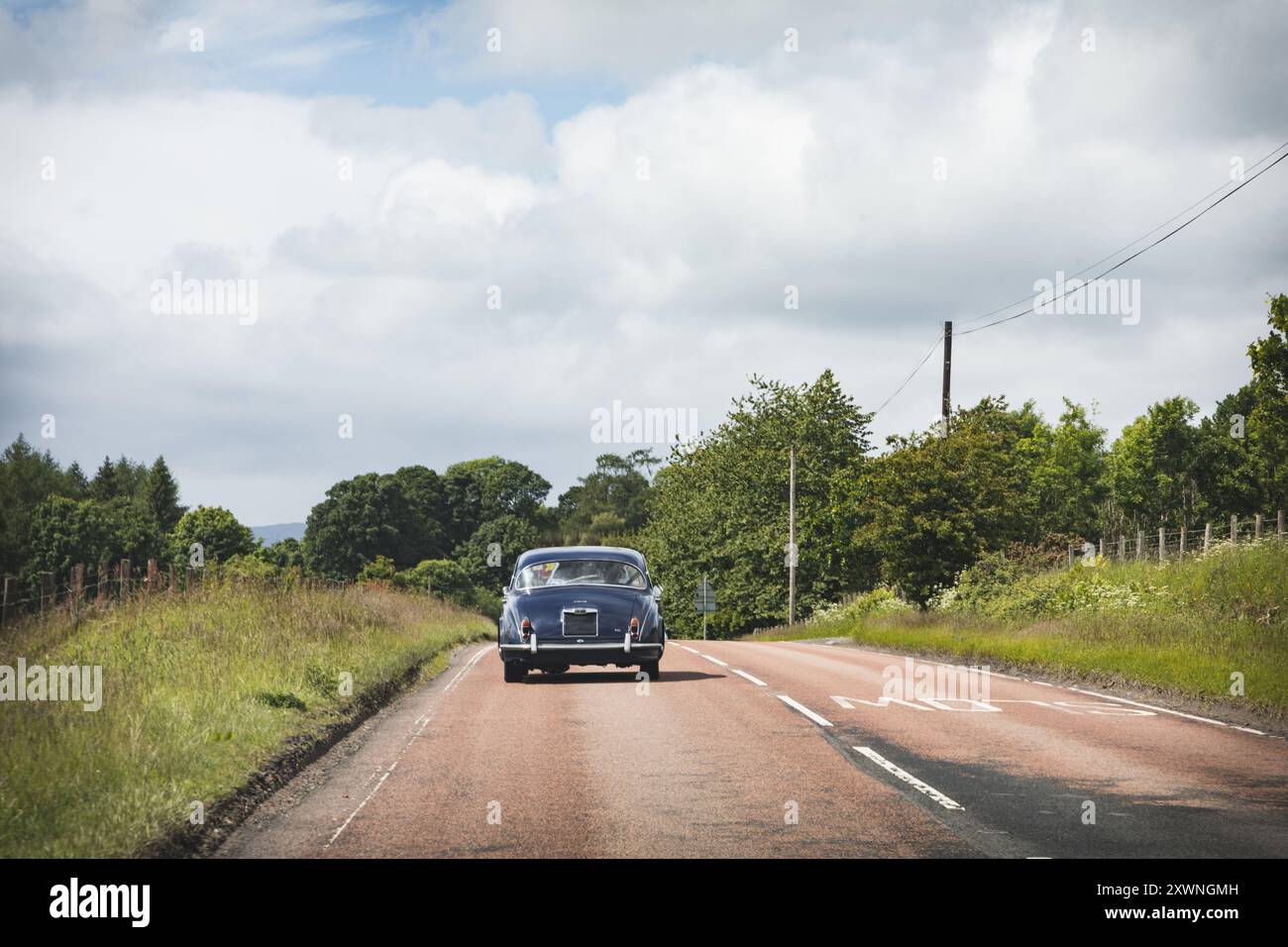 Ein Oldtimer fährt auf einer ruhigen, von Bäumen gesäumten Straße in der schottischen Landschaft unter einem bewölkten Himmel Stockfoto