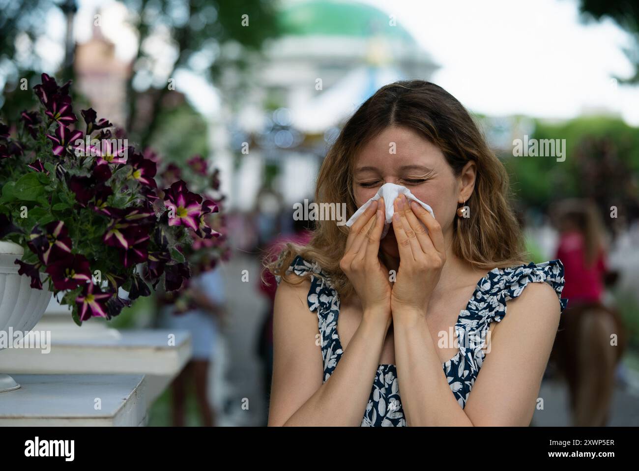 Frau niest in ein Taschentuch, während sie draußen in der Nähe von Blumen steht und saisonale Allergien in einer hellen, öffentlichen Umgebung erfährt. Stockfoto