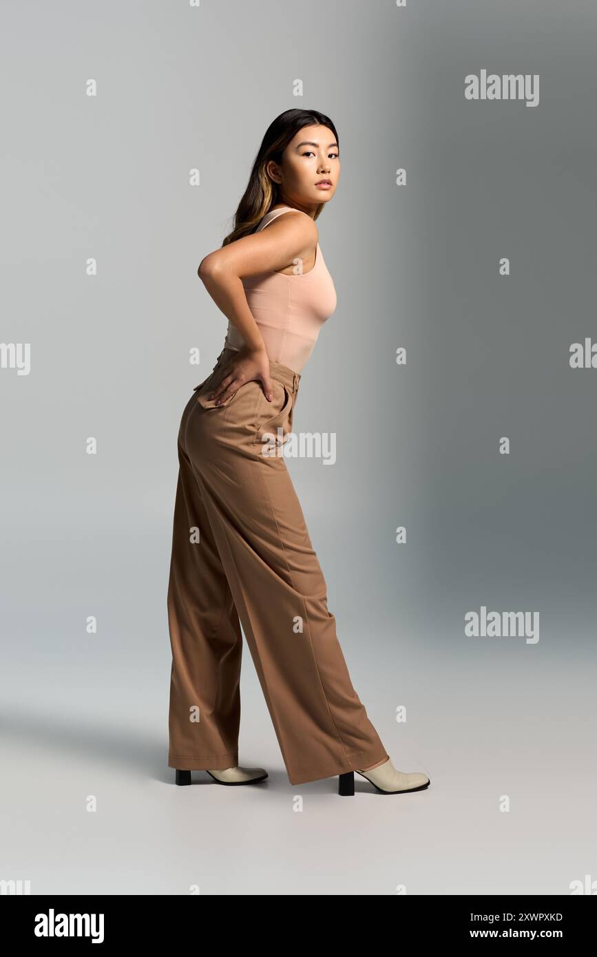 Eine Frau in stylischer brauner Hose und einem hellrosa Oberteil posiert selbstbewusst vor einem schlichten Hintergrund. Stockfoto