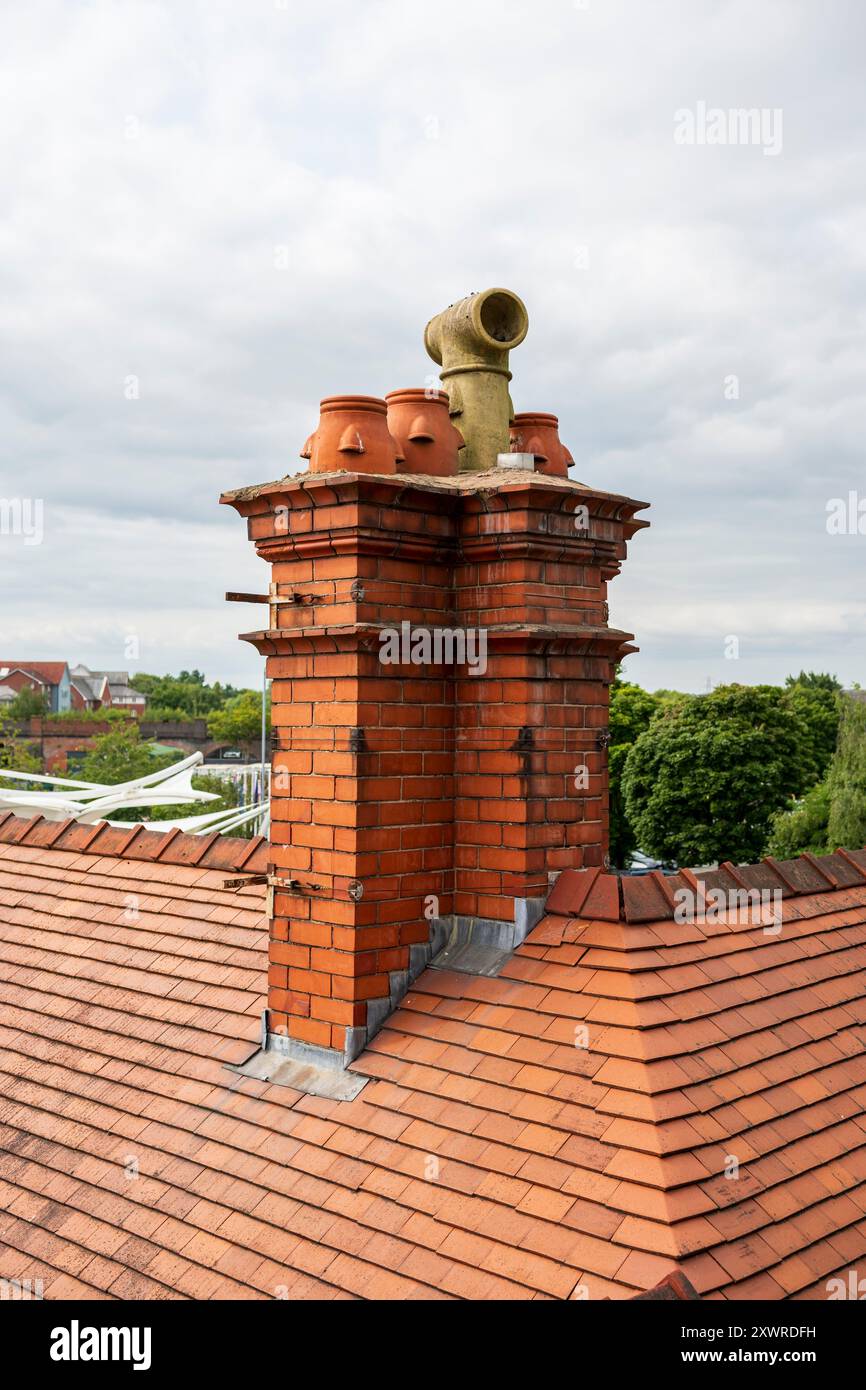 Alter roter Backsteinkamin auf einem gekachelten Dach, umgeben von üppigen grünen Bäumen und bewölktem Himmel im Hintergrund. UK Stockfoto