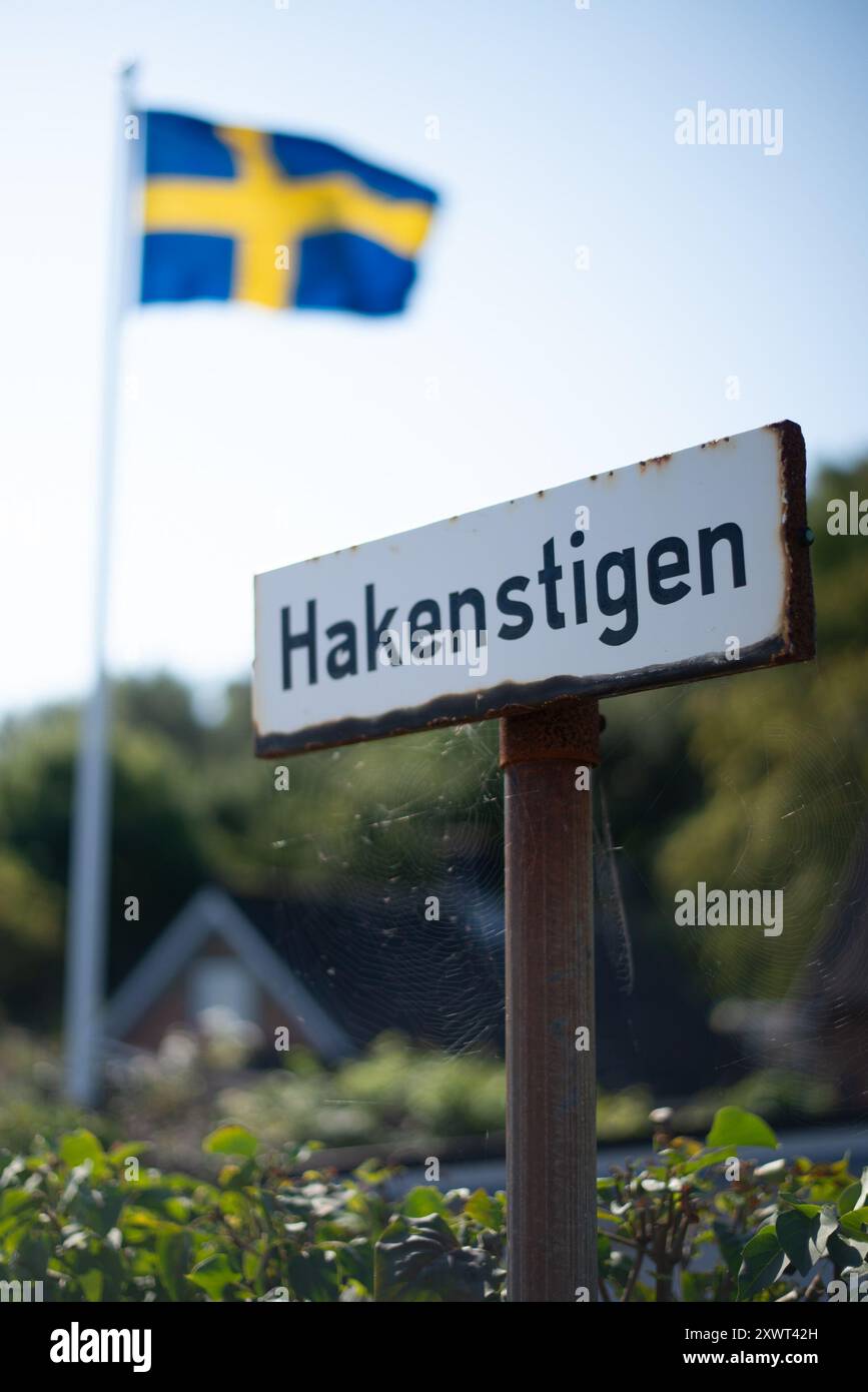 Ein Straßenschild mit der Aufschrift „Hakenstigen“ mit einer schwedischen Flagge, die an einem sonnigen Tag im Hintergrund winkt. Dieses Bild zeigt eine friedliche Nachbarschaftsszene in Schweden. Stockfoto