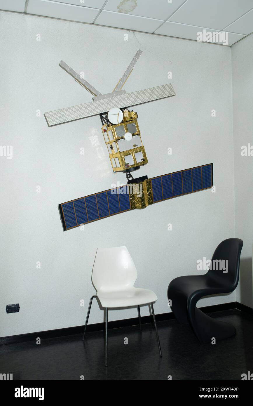 Ein detailliertes Modell eines Satelliten hängt an einer Wand mit zwei modernen Stühlen darunter. Diese Szene kann die Gegenüberstellung von technologischem Fortschritt und alltäglichem Leben symbolisieren. Stockfoto