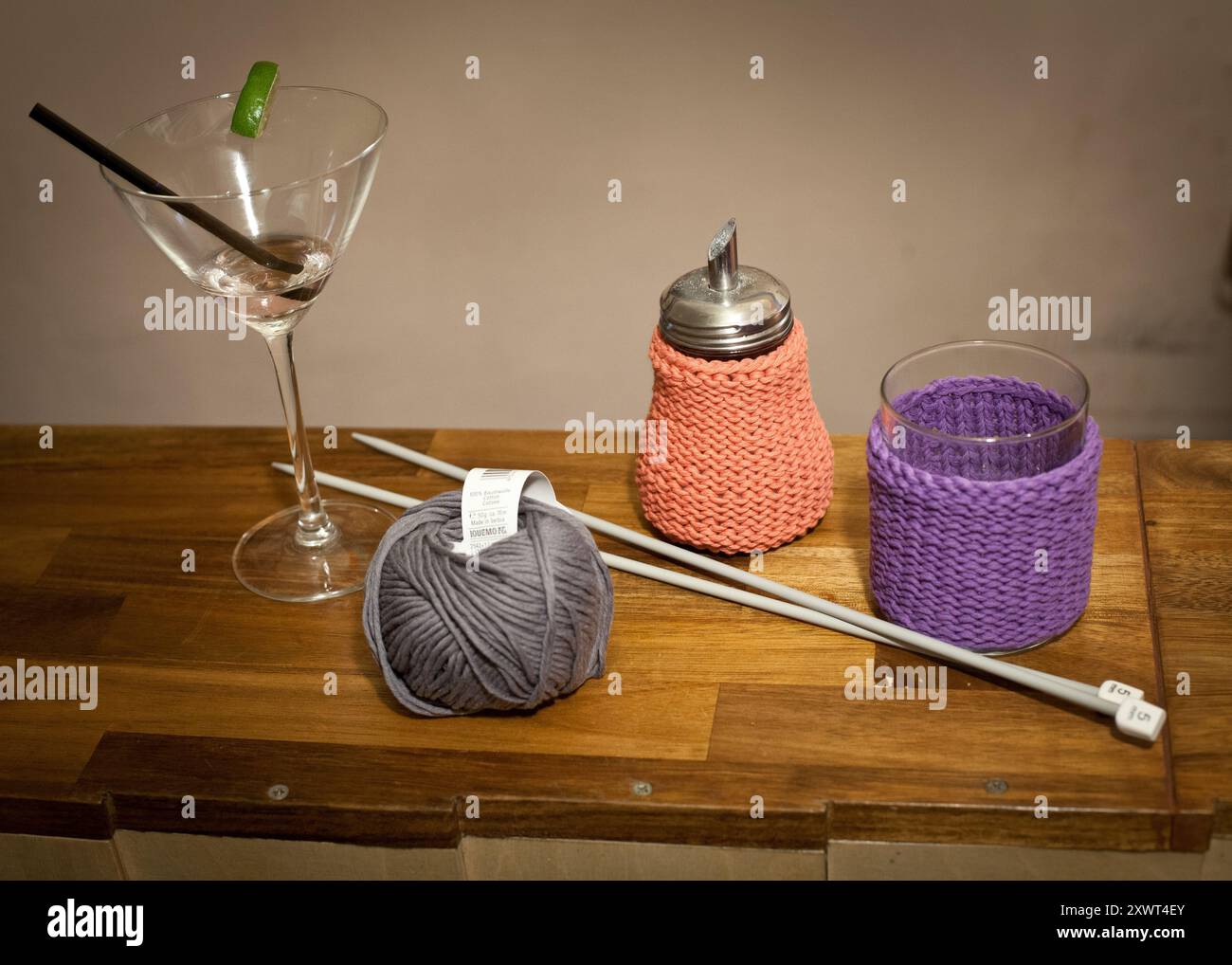 Ein Holztisch mit Martini-Glas mit Limettengarnitur, Stricknadeln, einem Garnknäuel und Behältern mit gestrickten Deckeln. Das Bild stellt eine Gegenüberstellung dar, die Freizeit und Basteln miteinander verbindet. Stockfoto