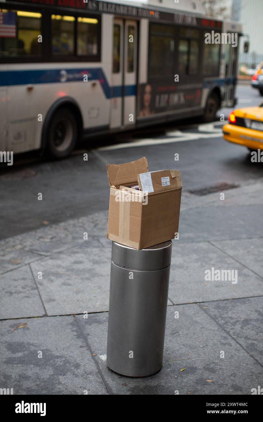 Ein verlassener Karton, der auf einem Straßenpfoller in einer städtischen Umgebung mit einem Bus und einem Taxi im Hintergrund sitzt. Dieses Bild kann Vernachlässigung oder vergessene Verantwortung symbolisieren. Stockfoto Ein verlassener Karton, der auf einem Straßenpfoller in einer städtischen Umgebung mit einem Bus und einem Taxi im Hintergrund sitzt. Dieses Bild kann Vernachlässigung oder vergessene Verantwortung symbolisieren. Stockfoto