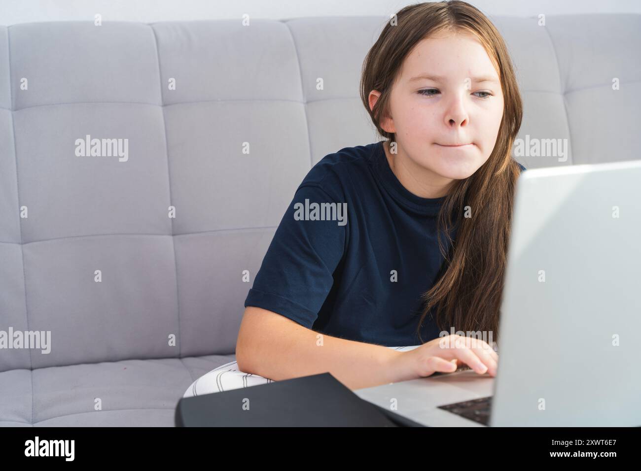 Kleines Mädchen im Teenageralter, das zu Hause auf einem grauen Sofa in blauem T-Shirt sitzt. Das Konzept der Online-Kommunikation und des Lernens des Codierens. Hochwertige Pho Stockfoto