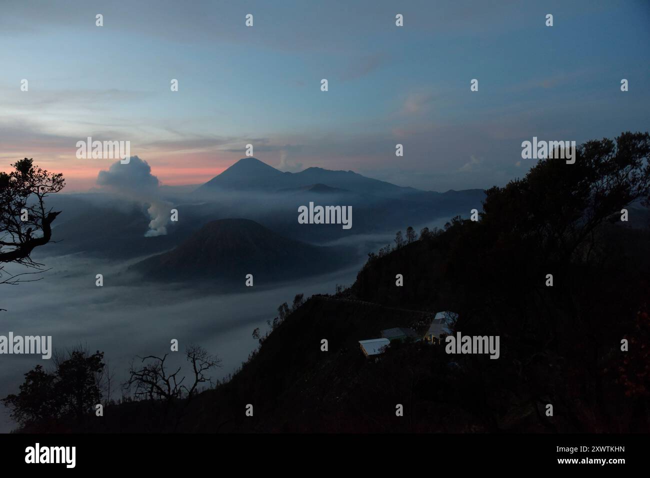 Frühnebel bei Sonnenaufgang in der Tengger-Caldera. Links ist der Bromo rauchender Krater / Höhe: 2,329 Meter und rechts vorne der Batok zu sehen, im Hintergrund der Vulkan Semeru mit 3676 m Höhe der höchste Berg der indonesischen Insel Java. Der Nationalpark Bromo-Tengger-Semeru besteht seit 1982. Der mittlere Teil des Namens verweist auf das hier lebende Volk der Tengger. Zum Nationalpark gehören der 3676 m hohe Stratovulkan Semeru, der höchste Berg der Insel, sowie vier in der Tengger-Caldera liegende Vulkane, deren Bekanntester der Bromo ist. Die Caldera wird vom Tengger-Sandmeer malaiisch Stockfoto