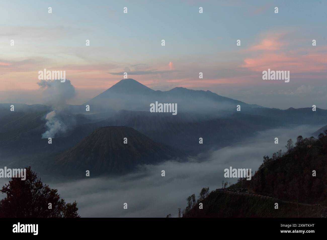 Frühnebel bei Sonnenaufgang in der Tengger-Caldera. Links ist der Bromo rauchender Krater / Höhe: 2,329 Meter und rechts vorne der Batok zu sehen, im Hintergrund der Vulkan Semeru mit 3676 m Höhe der höchste Berg der indonesischen Insel Java. Der Nationalpark Bromo-Tengger-Semeru besteht seit 1982. Der mittlere Teil des Namens verweist auf das hier lebende Volk der Tengger. Zum Nationalpark gehören der 3676 m hohe Stratovulkan Semeru, der höchste Berg der Insel, sowie vier in der Tengger-Caldera liegende Vulkane, deren Bekanntester der Bromo ist. Die Caldera wird vom Tengger-Sandmeer malaiisch Stockfoto