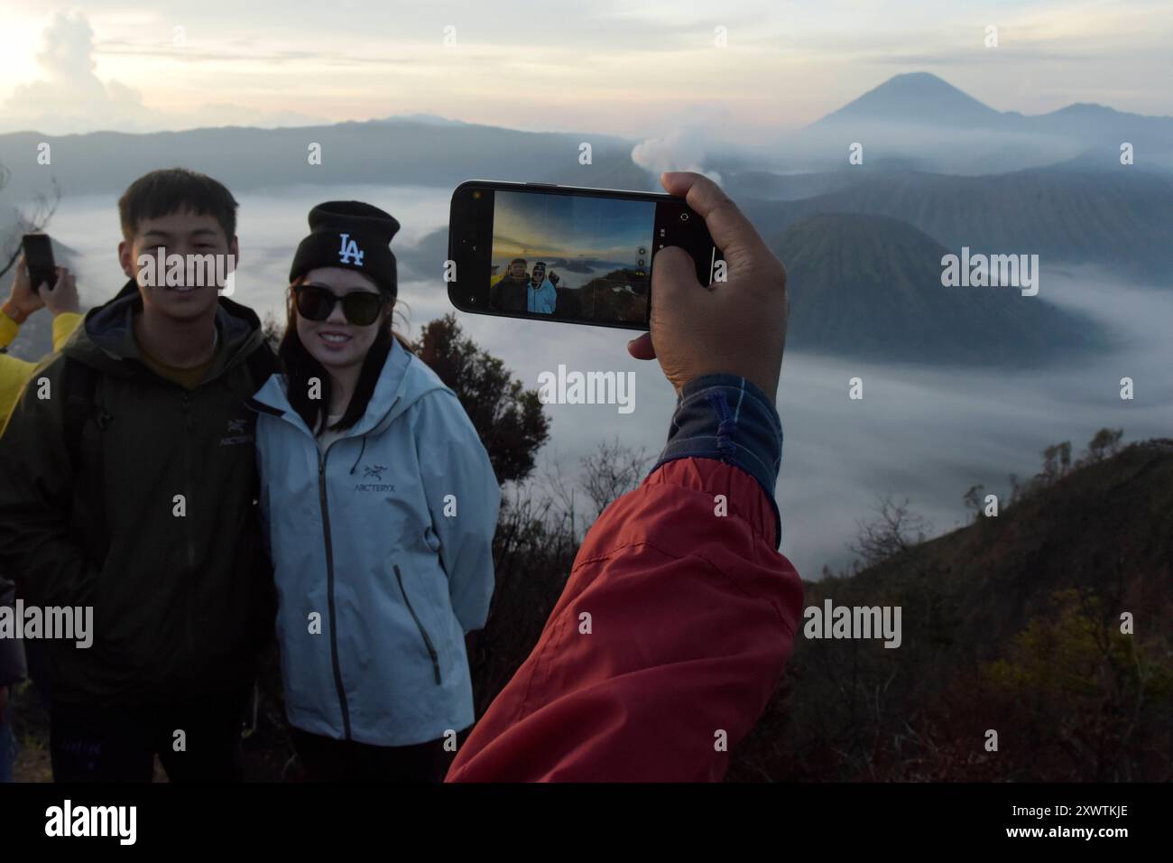 Ein Pärchen lässt sich bei Sonnenaufgang vor dem Tengger-Caldera fotografieren. Im Hintergrund ist der Vulkan Semeru, mit 3676 m Höhe der höchste Berg der indonesischen Insel Java, zu sehen. Der Nationalpark Bromo-Tengger-Semeru besteht seit 1982. Der mittlere Teil des Namens verweist auf das hier lebende Volk der Tengger. Zum Nationalpark gehören der 3676 m hohe Stratovulkan Semeru, der höchste Berg der Insel, sowie vier in der Tengger-Caldera liegende Vulkane, deren Bekanntester der Bromo ist. Die Caldera wird vom Tengger-Sandmeer malaiisch: Laut Pasir Tengger ausgefüllt. Vier gesehen und etwa Stockfoto