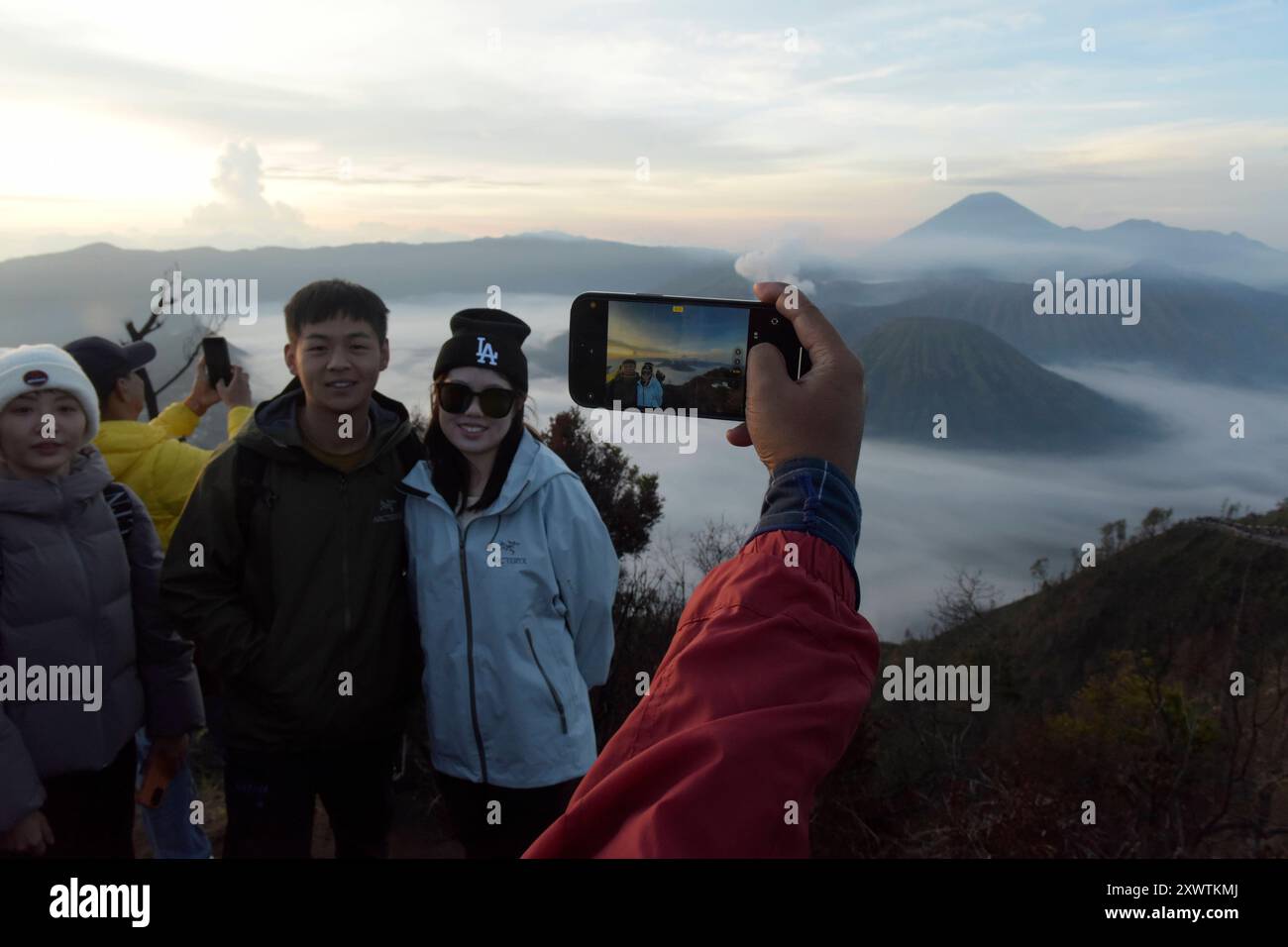 Ein Pärchen lässt sich bei Sonnenaufgang vor dem Tengger-Caldera fotografieren. Im Hintergrund ist der Vulkan Semeru, mit 3676 m Höhe der höchste Berg der indonesischen Insel Java, zu sehen. Der Nationalpark Bromo-Tengger-Semeru besteht seit 1982. Der mittlere Teil des Namens verweist auf das hier lebende Volk der Tengger. Zum Nationalpark gehören der 3676 m hohe Stratovulkan Semeru, der höchste Berg der Insel, sowie vier in der Tengger-Caldera liegende Vulkane, deren Bekanntester der Bromo ist. Die Caldera wird vom Tengger-Sandmeer malaiisch: Laut Pasir Tengger ausgefüllt. Vier gesehen und etwa Stockfoto