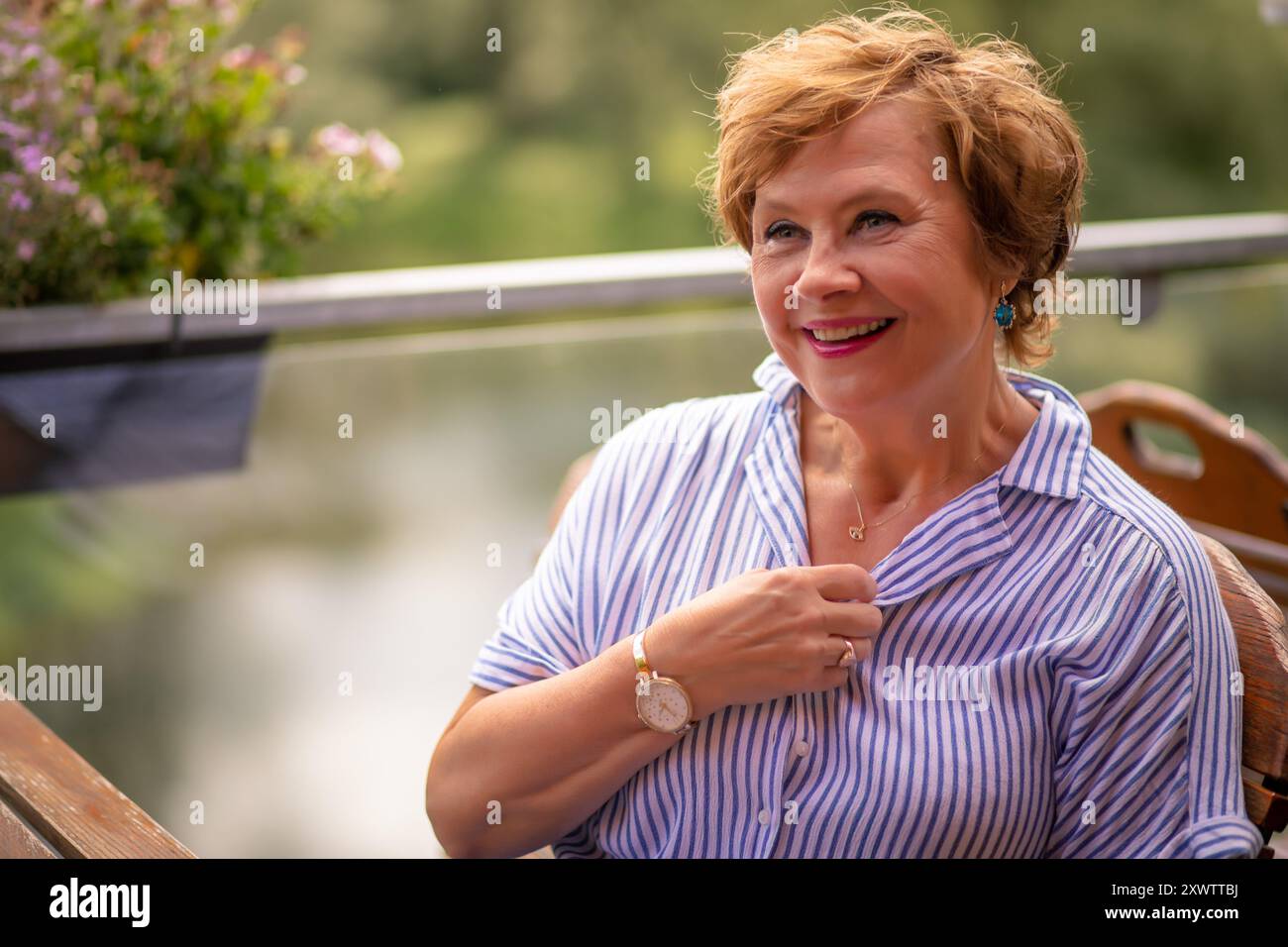 Eine fröhliche Frau sitzt in einem gestreiften Hemd auf einer Terrasse am See und genießt einen warmen Tag und die malerische Aussicht. Stockfoto