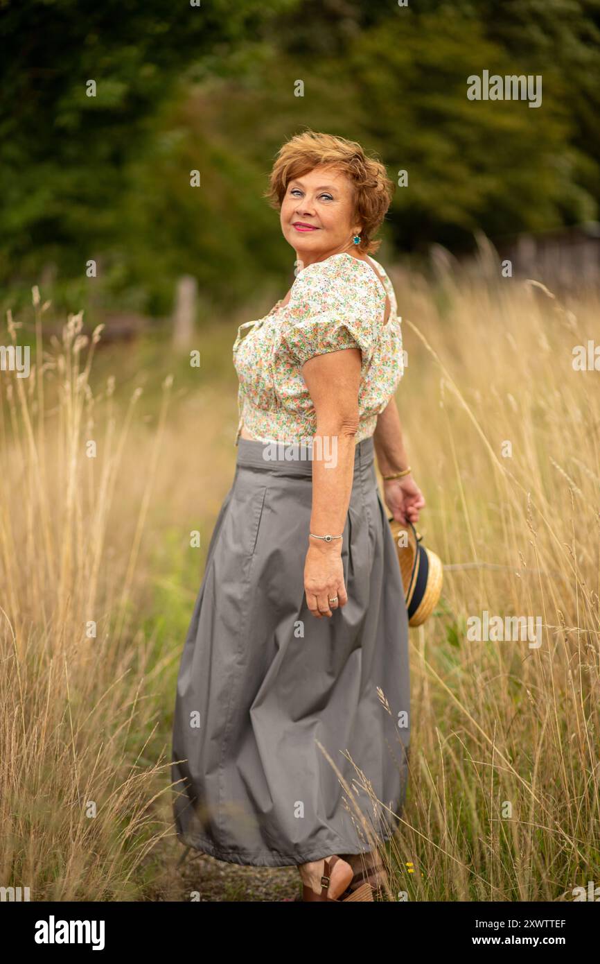 Eine ältere Frau schlendert durch hohes Gras in einer Blumenkohle und einem langen Rock und genießt einen sonnigen Tag in einer ruhigen Landschaft. Stockfoto