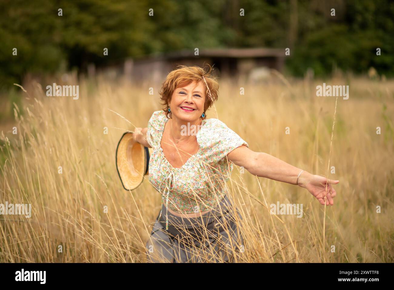 Eine Frau mit einem strahlenden Lächeln wirbelt in einem Feld aus hohem Gras, hält einen Sonnenhut und genießt einen sonnigen Moment in der Natur. Stockfoto