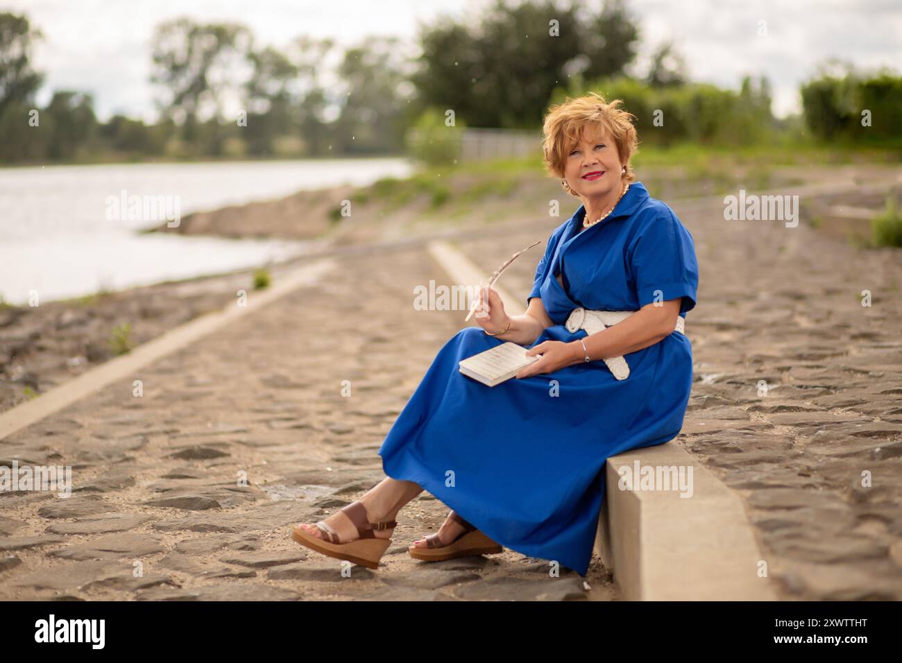 Eine Frau genießt einen friedlichen Moment am Wasser und schreibt Gedanken in ihr Notizbuch, während sie auf einem Steinweg sitzt. Stockfoto