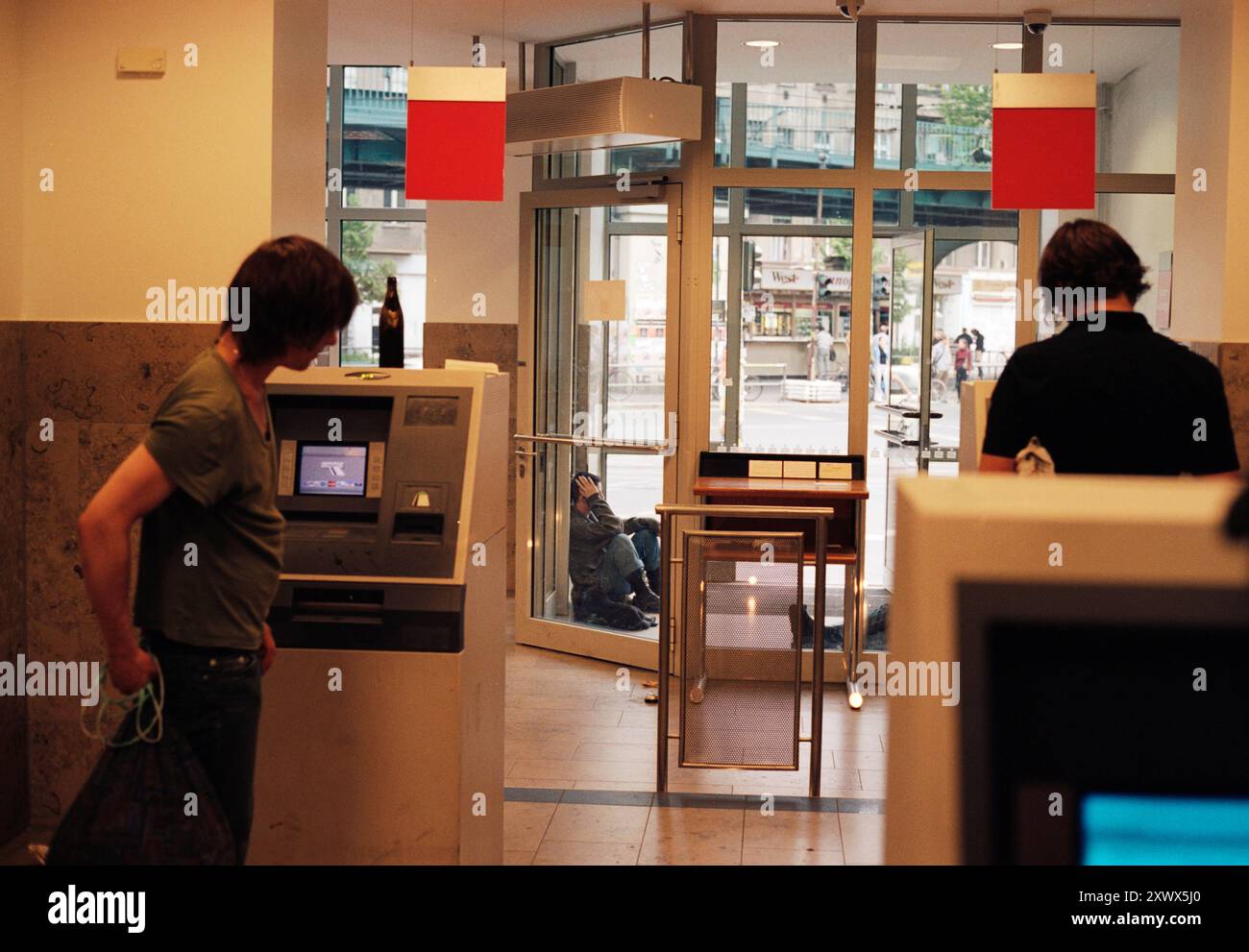 Eine Alltagsszene in einer Berliner Bankfiliale mit Leuten, die Geldautomaten benutzen. Das Bild fängt die tägliche Routine ein und deutet auf persönliche Momente und finanzielle Transaktionen hin, mit einem Kontrast zu den Straßen draußen. Stockfoto