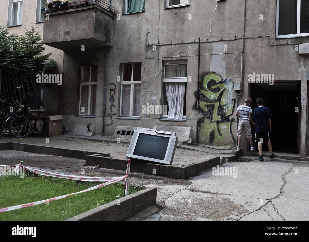 Ein alter, kaputter Fernseher liegt in einem Berliner Innenhof, umgeben von Graffiti-bedeckten Wänden und neugierigen Kindern. Die Szene spiegelt urbanen Verfall und Erkundung im Stadtteil Neukölln wider, mit einem Gefühl von Nostalgie und Wandel. Stockfoto
