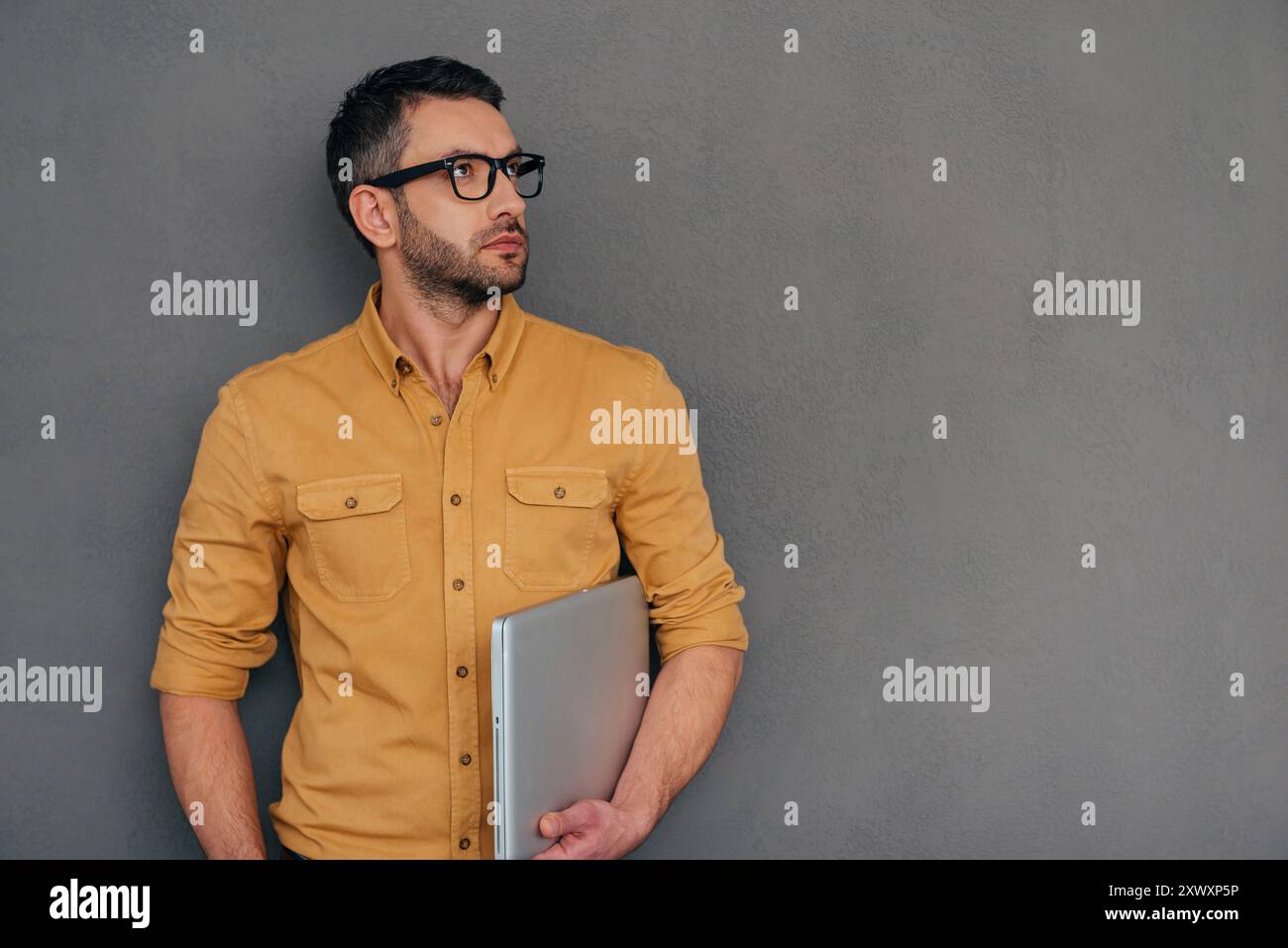 In geschäftlichen Gedanken verloren. Ein durchdachter reifer Mann, der ein Notebook trägt und wegblickt, während er vor grauem Hintergrund steht Stockfoto