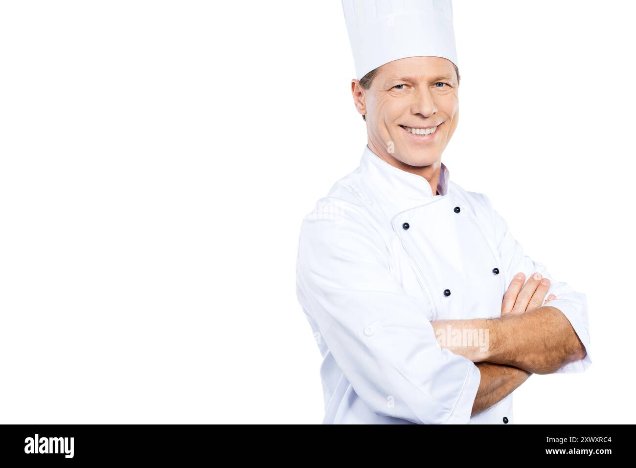 Kochen ist meine Leidenschaft. Der selbstbewusste, reife Koch in weißer Uniform hält die Arme gekreuzt und lächelt, während er vor weißem Hintergrund steht Stockfoto