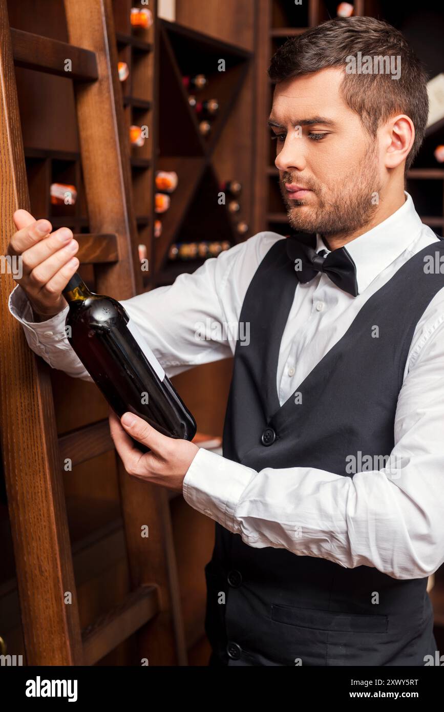 Dieser Wein ist perfekt. Selbstbewusster männlicher Sommelier, der die Weinflasche untersucht, während er in der Nähe des Weinregals steht Stockfoto