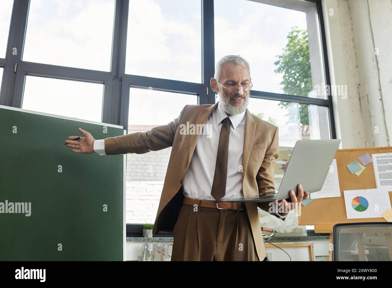 Ein bärtiger, reifer Lehrer in einem Klassenzimmer unterrichtet Schüler online über seinen Laptop. Stockfoto