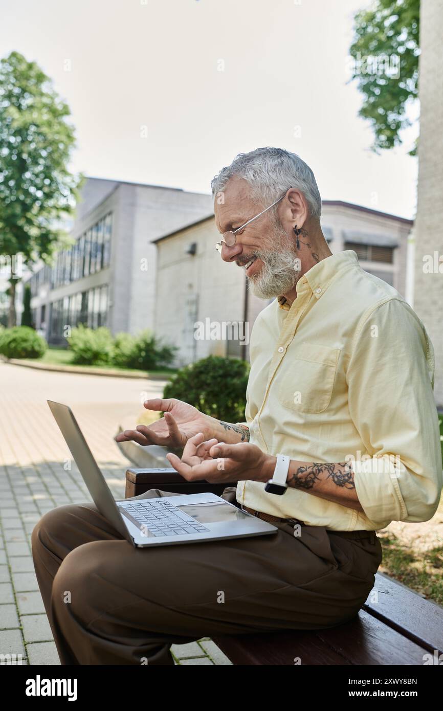 Ein reifer Mann mit Bart sitzt auf einer Bank und lächelt, während er seinen Laptop benutzt. Er trägt ein hellgelbes Hemd und hat ein Tattoo am Arm. Stockfoto