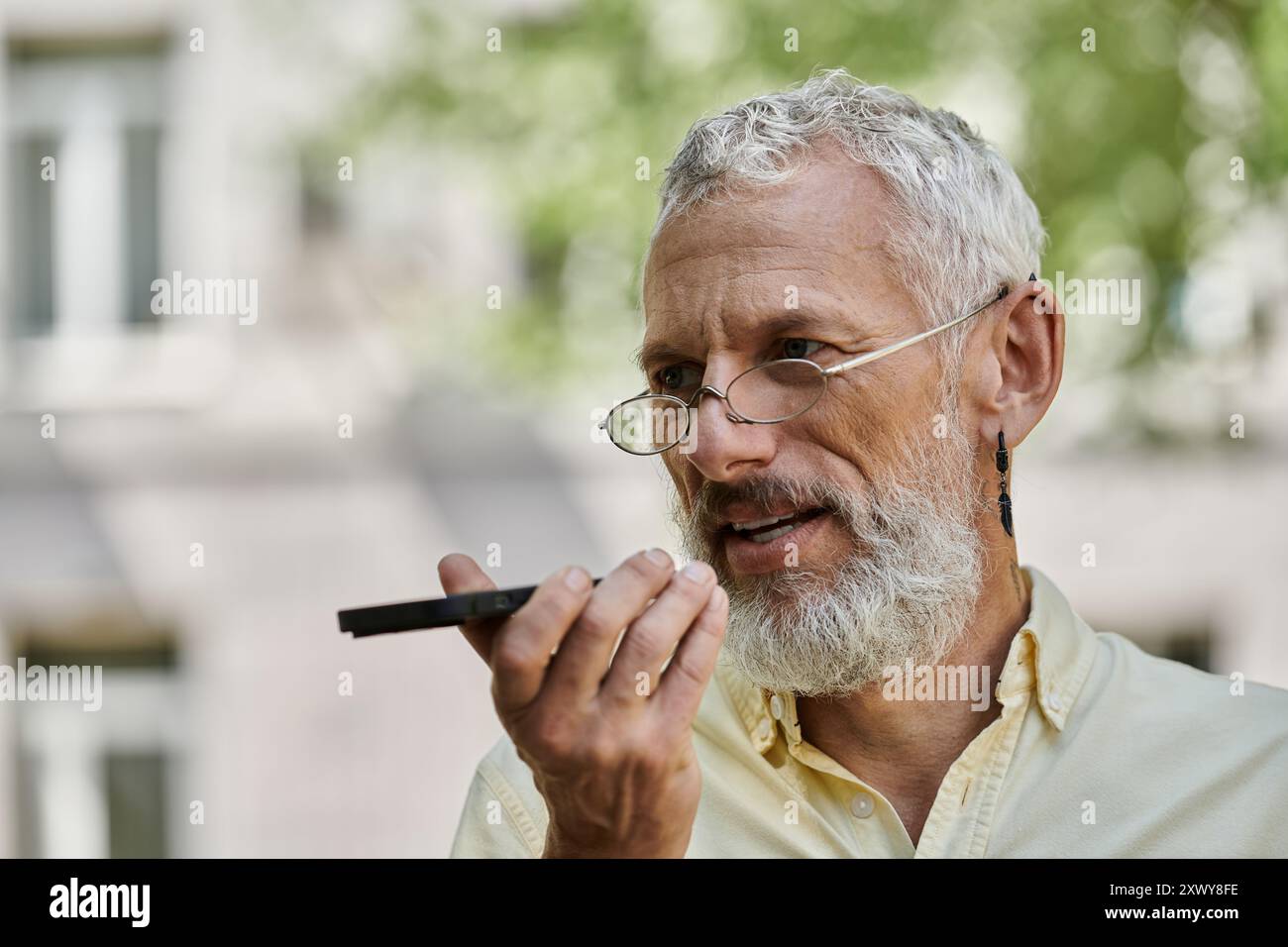 Ein Mann mittleren Alters mit Bart und Brille spricht in ein Aufnahmegerät, während er vor einem modernen Gebäude steht. Stockfoto