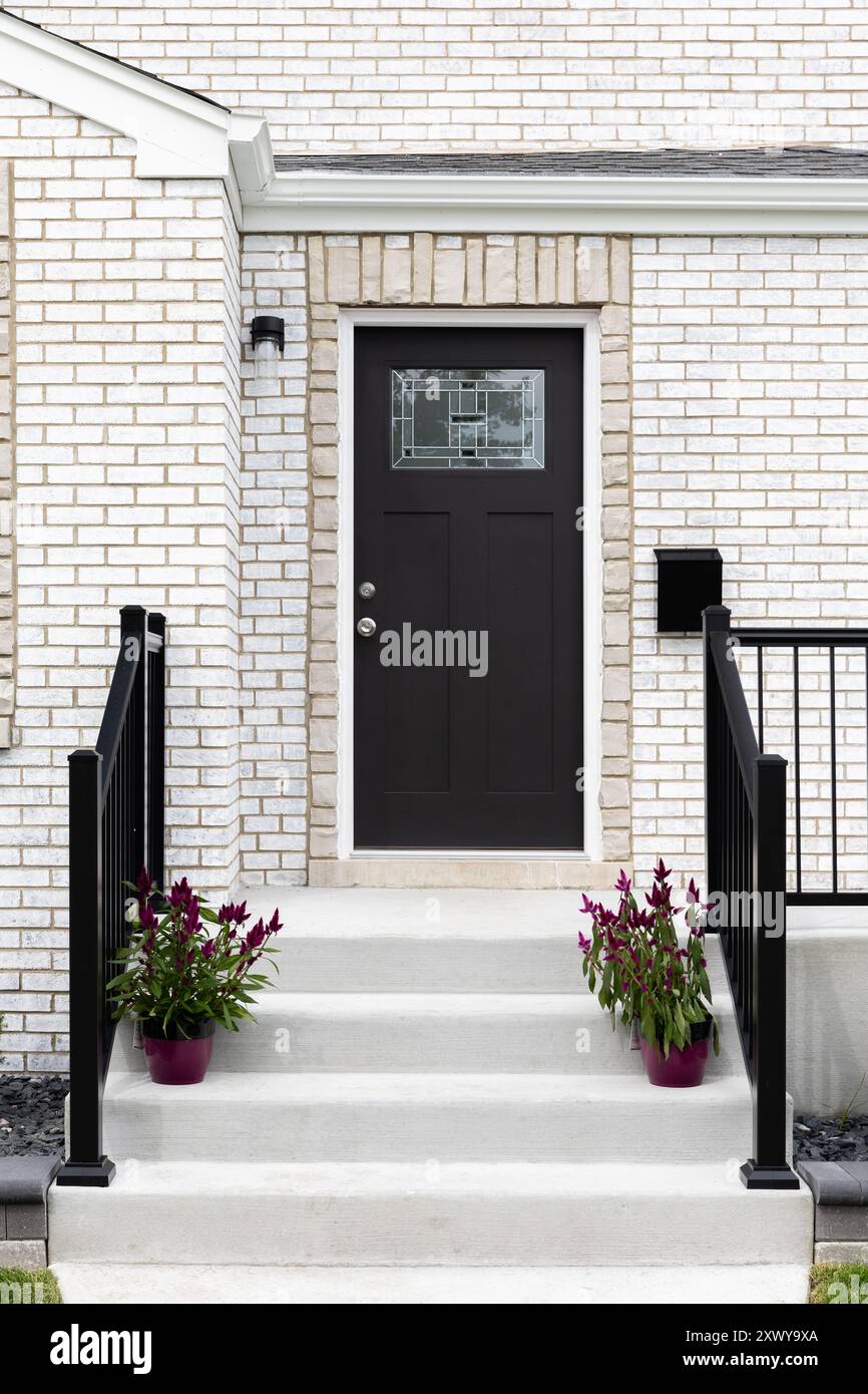 Eine Eingangstür in einem weißen Backsteinhaus mit Betontreppen und schwarzem Geländer führt zu einer schwarzen Eingangstür mit Mosaikfenster. Stockfoto