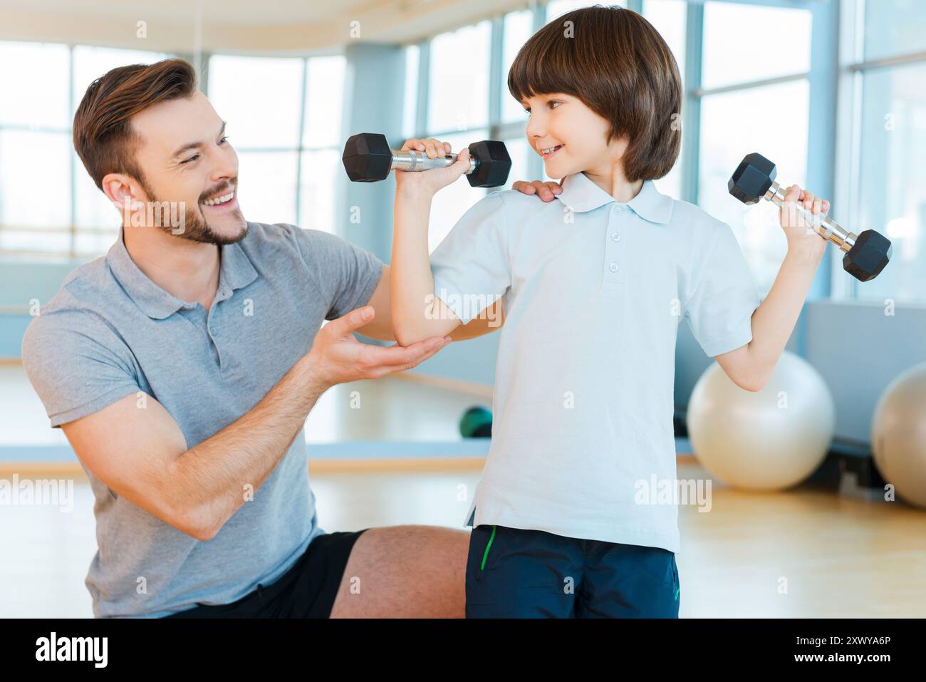 Bereit, stark und gesund zu sein. Glücklicher Vater hilft seiner Sonne mit Gewichtstraining, während beide im Health Club stehen Stockfoto