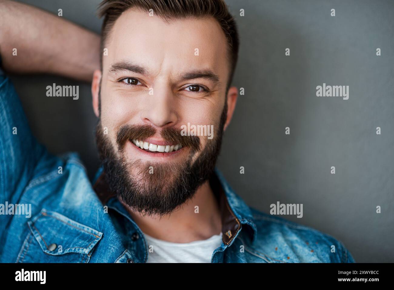 Glück kommt nie aus der Mode. Fröhlicher junger Mann, der Hand hinter Kopf hält und in die Kamera lächelt, während er vor grauem Hintergrund steht Stockfoto