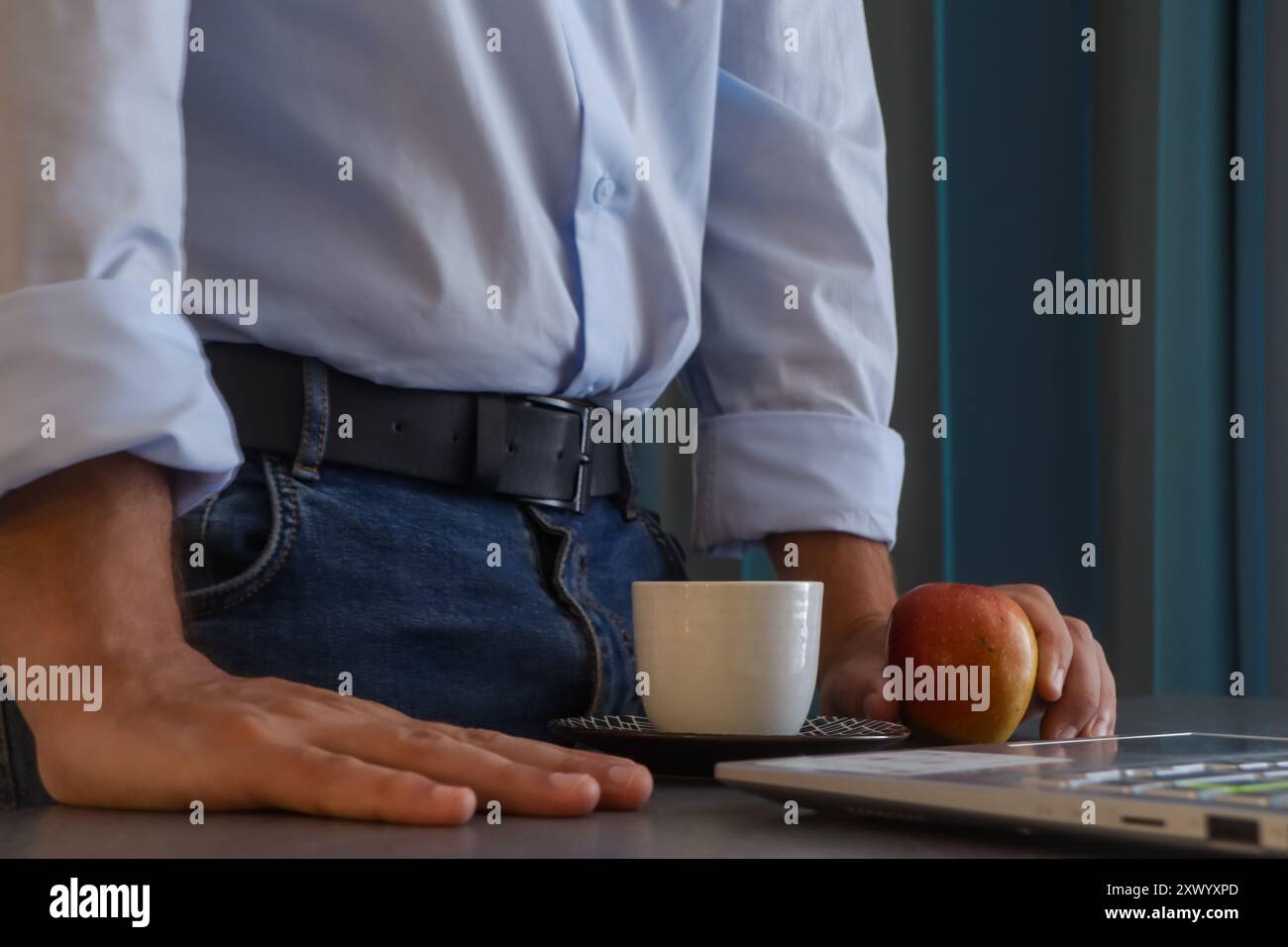 Der Mann steht auf, um während seines Arbeitstages eine Pause einzulegen, und er nimmt eine Tasse Kaffee und ein Stück Obst vor seinem Laptop Stockfoto