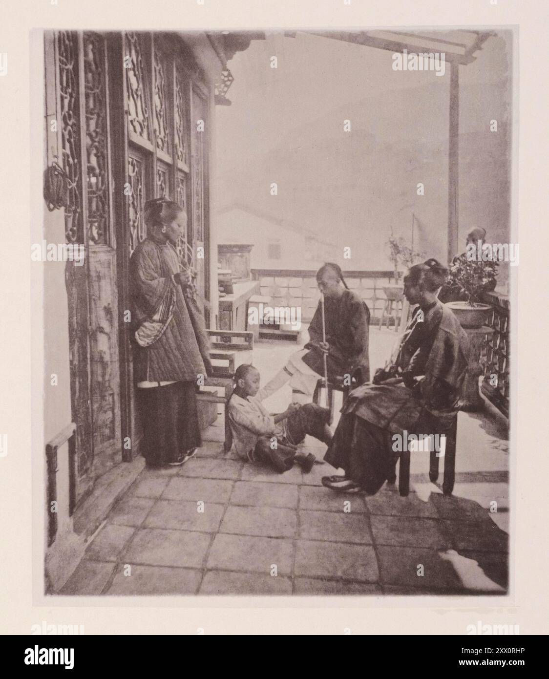 Vintage-Foto des Alltags in Hongkong des 19. Jahrhunderts. Treffpunkt nach dem Abendessen. Illustrations of Qing China, 1875 Dies ist eine Zusammenkunft nach dem Abendessen auf der Veranda eines Chinamannshauses. Der gesamte Hauskreis raucht Tabak, aber ihre Pfeifen unterscheiden sich von unseren. Die alte Frau und ihre Tochter benutzen eine Pfeife, die der "Shisha" ähnelt und ein kleines Fach mit Wasser hat, um den Rauch zu kühlen und zu reinigen. Paterfamilias streichelt und saugt das Ende eines scheinbar sehr beeindruckenden Gehstockes; aber es ist in Wirklichkeit seine Lieblingspfeife, die eine geliebte Geschichte hat Stockfoto