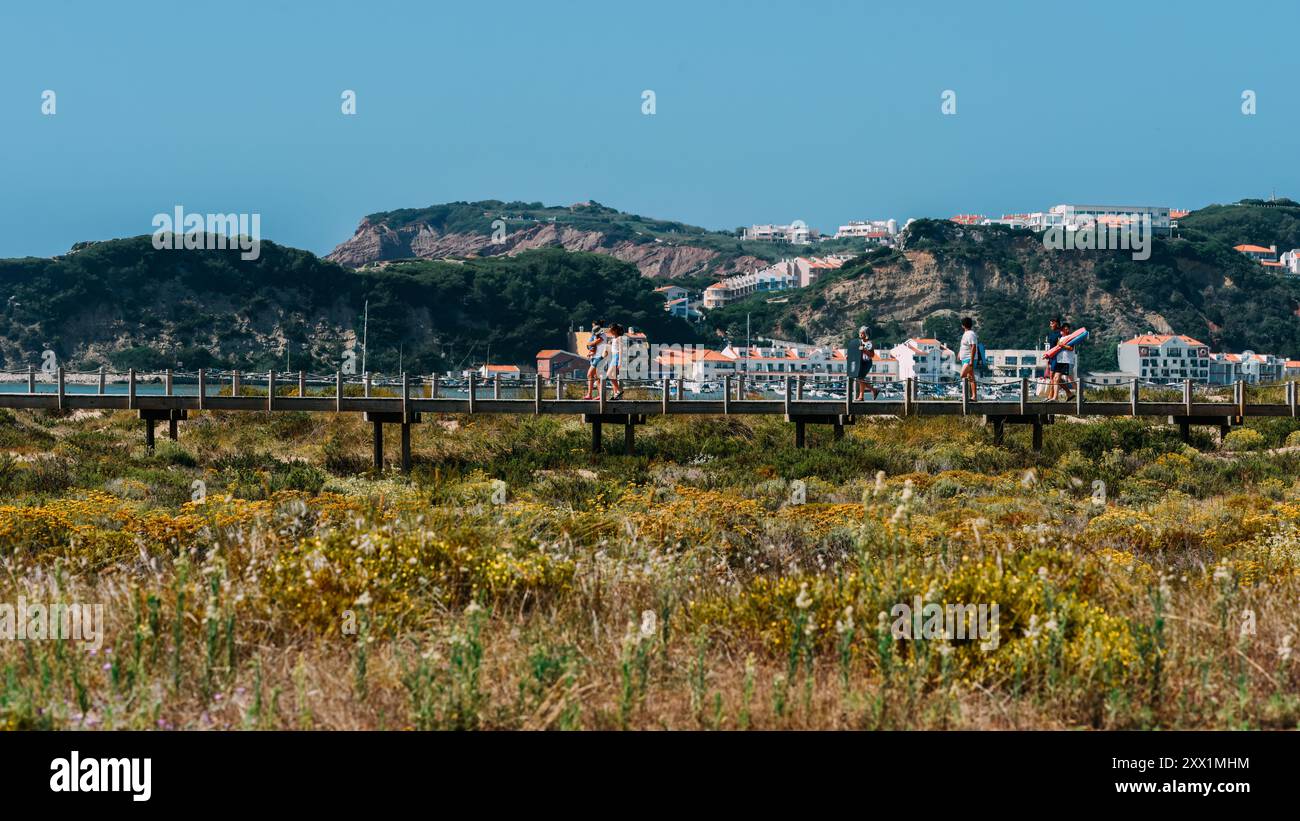 Besucher schlendern entlang des hölzernen Spaziergangs in Sao Martinho do Porto, umgeben von lebendigen Wildblumen und atemberaubendem Blick auf die Küste, Oeste, Portugal Stockfoto