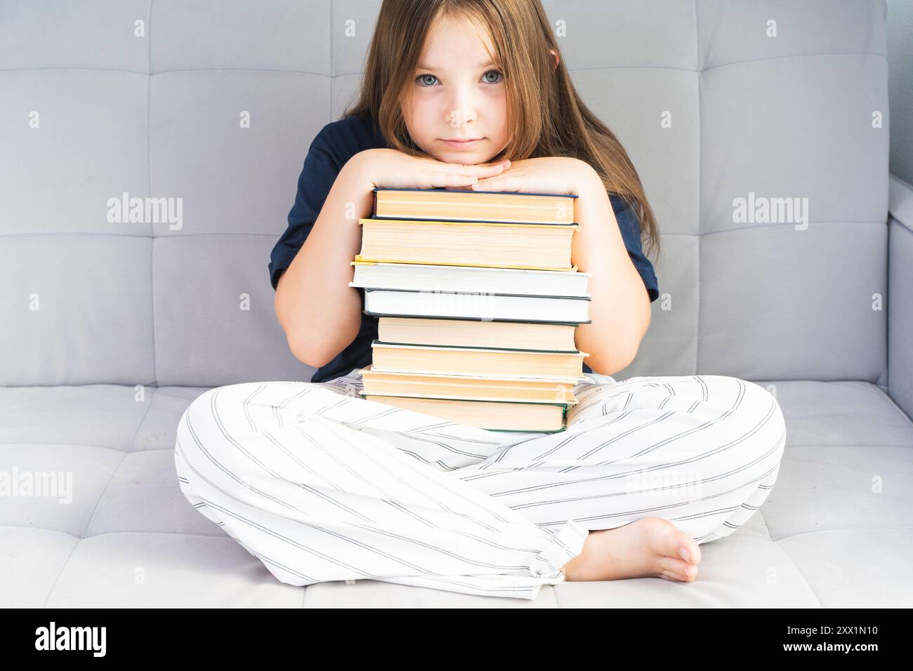 Kleines Mädchen, das einen Stapel Bücher hält, Hände und Kopf darauf legt, auf einem grauen Sofa sitzt. Hochwertige Fotos Stockfoto