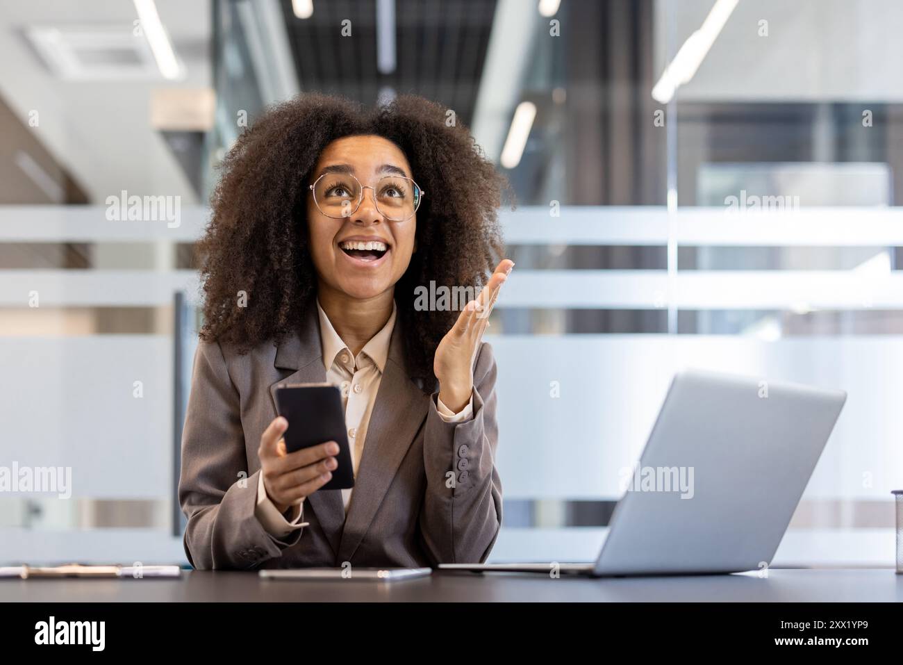 Glückliche junge afroamerikanische Geschäftsfrau in Anzug und Brille freut sich, Nachrichten am Telefon zu erhalten und lächelt dank erhobener Hände hoch. Stockfoto